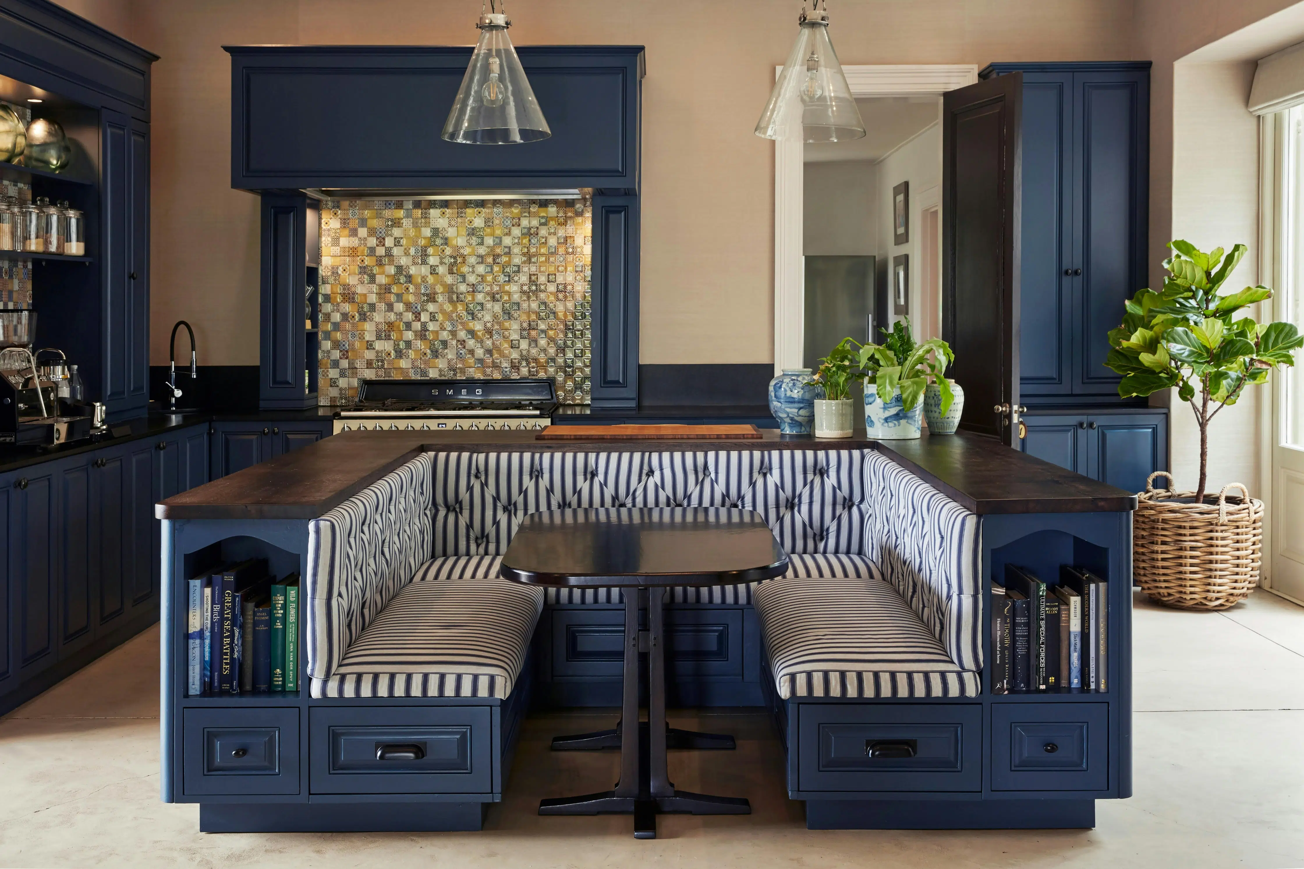 A breakfast nook with striped, upholstered seating and a blue wooden frame stands at the centre of a large kitchen. In the sides of the wooden breakfast nook are built-in drawers and shelves holding cookbooks. A cluster of potted plants flourishes in the background, a fiddle-leaf fig stands in a woven basket on the far right, and a decorative tile splashback is seen in the background, in what appears to be an alcove for the stove.