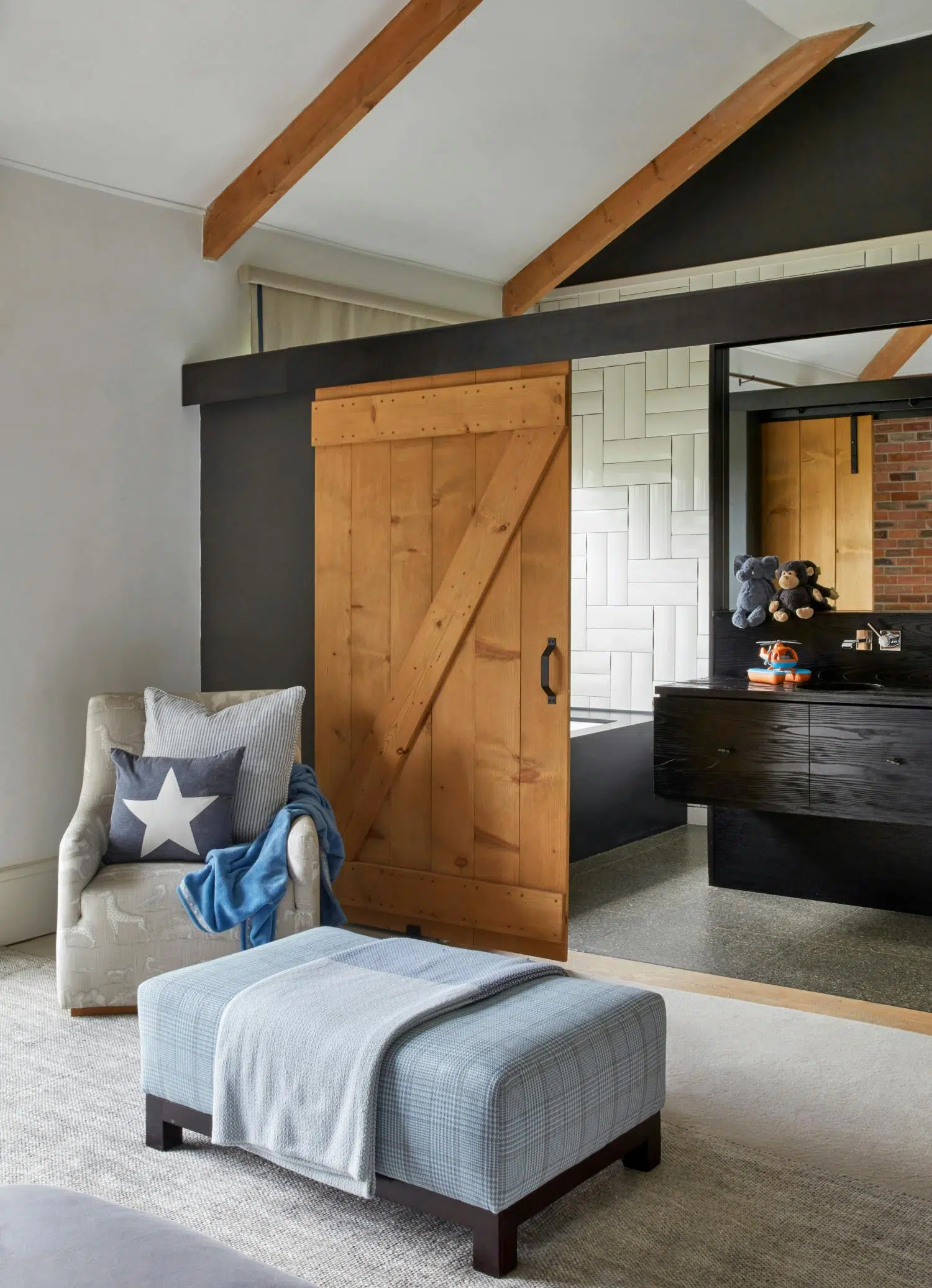 A blue plaid ottoman and an armchair in beige with giraffe patterns it in front of an open, wooden door. Both bold blankets, and the couch holds two cushions, one with a star motif. Through the door seems to be a bold, minimal bathroom with a child's helicopter toy and two plush animals on the vanity.