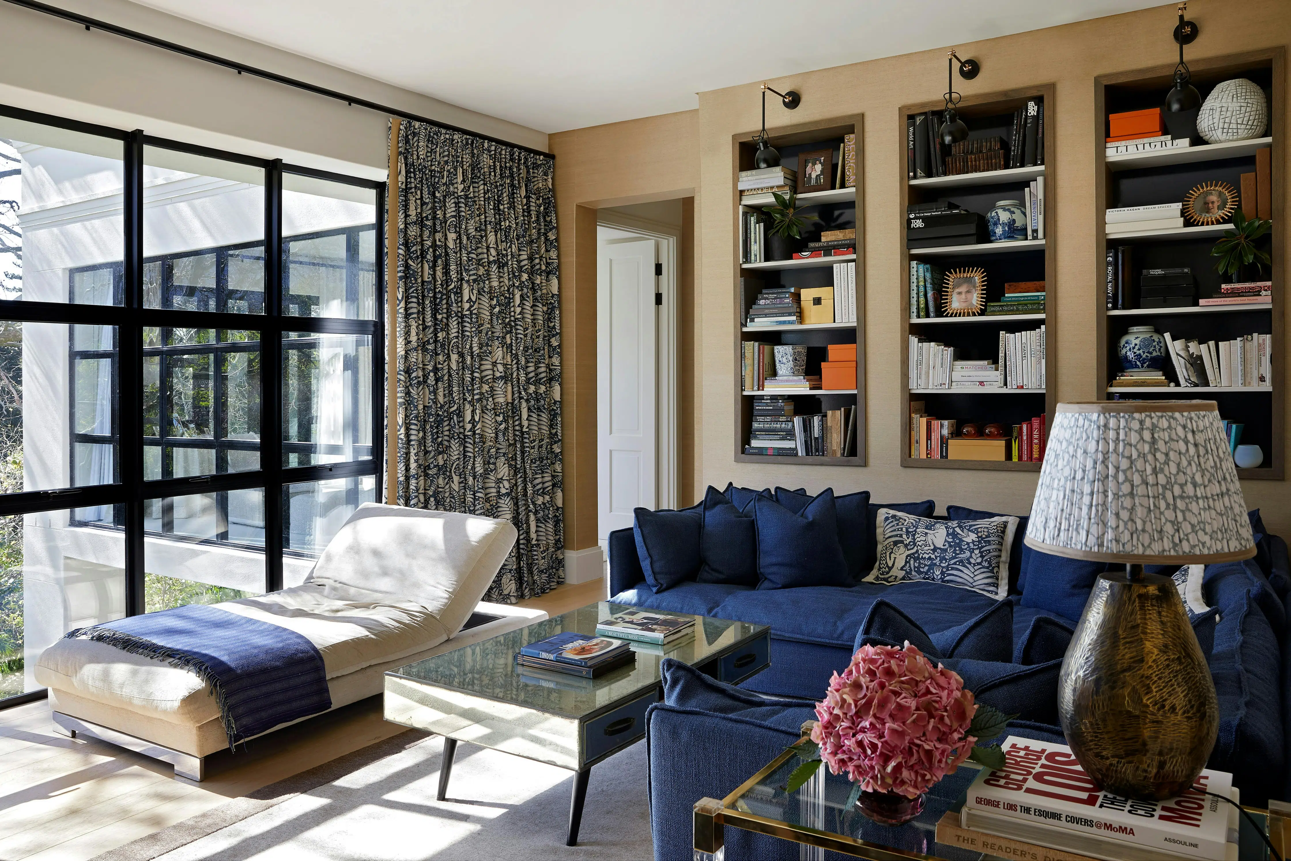 Shelving in the background that features books and decorative accents, with a daybed near large windows on the left, a coffee table in the middle and a blue sofa to the right.