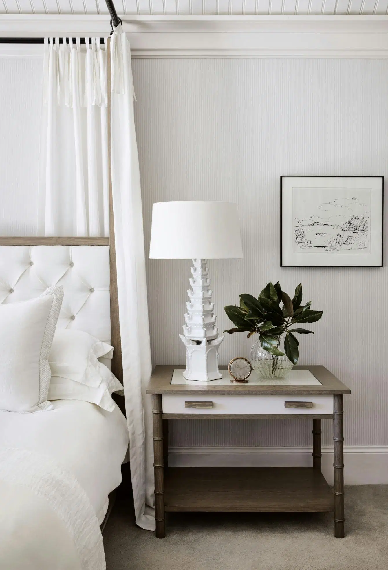 A closeup of a side table shows a clock, contemporary lamp and glass vase holding green foliage. On the wall is a line drawing, and the wallpaper and bed linen are white.