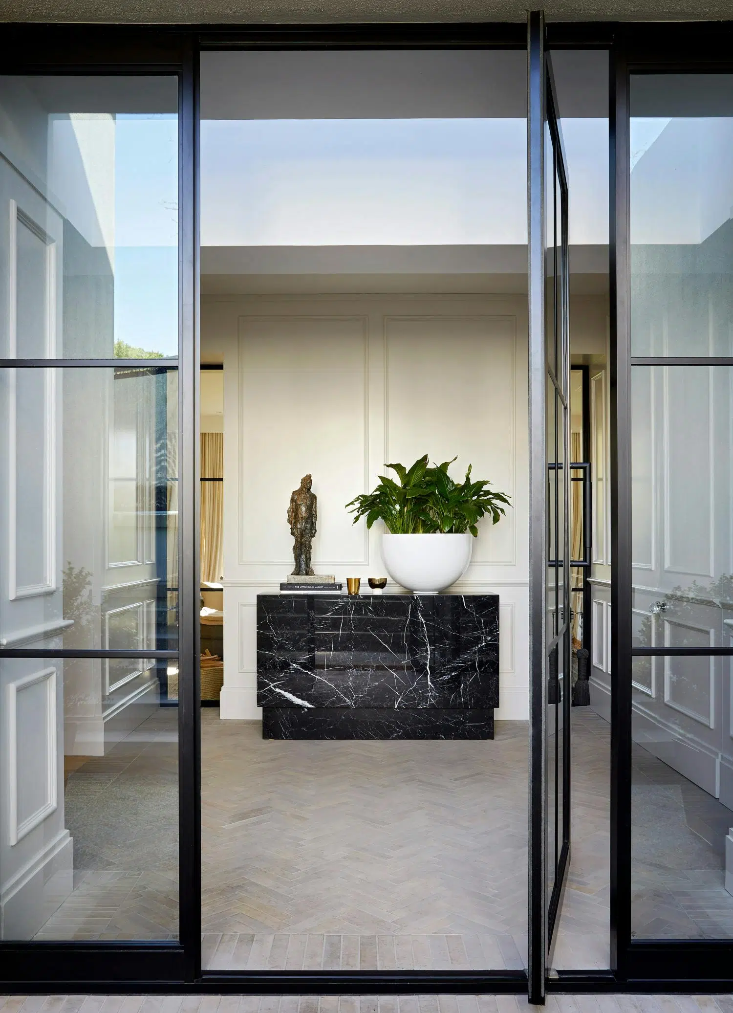 View of a front entrance to a home with with a large potted plant and a sculpture of a man on top of a marble console table in the centre.