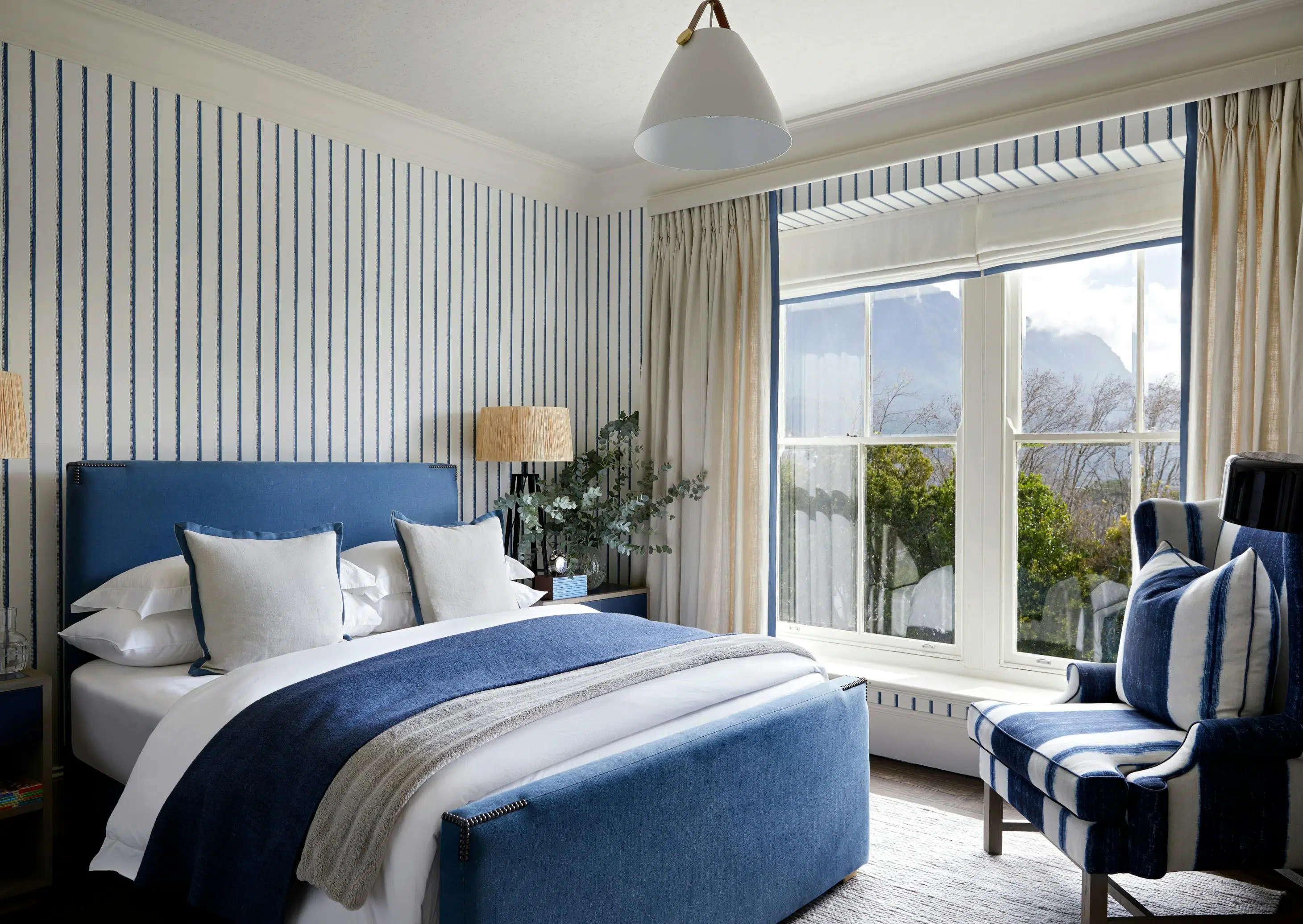 A contemporary bedroom with blue and white striped wallpaper, a navy blue bed layered in blue, white and grey bedding and an armchair in the corner next to a window with a view of trees.