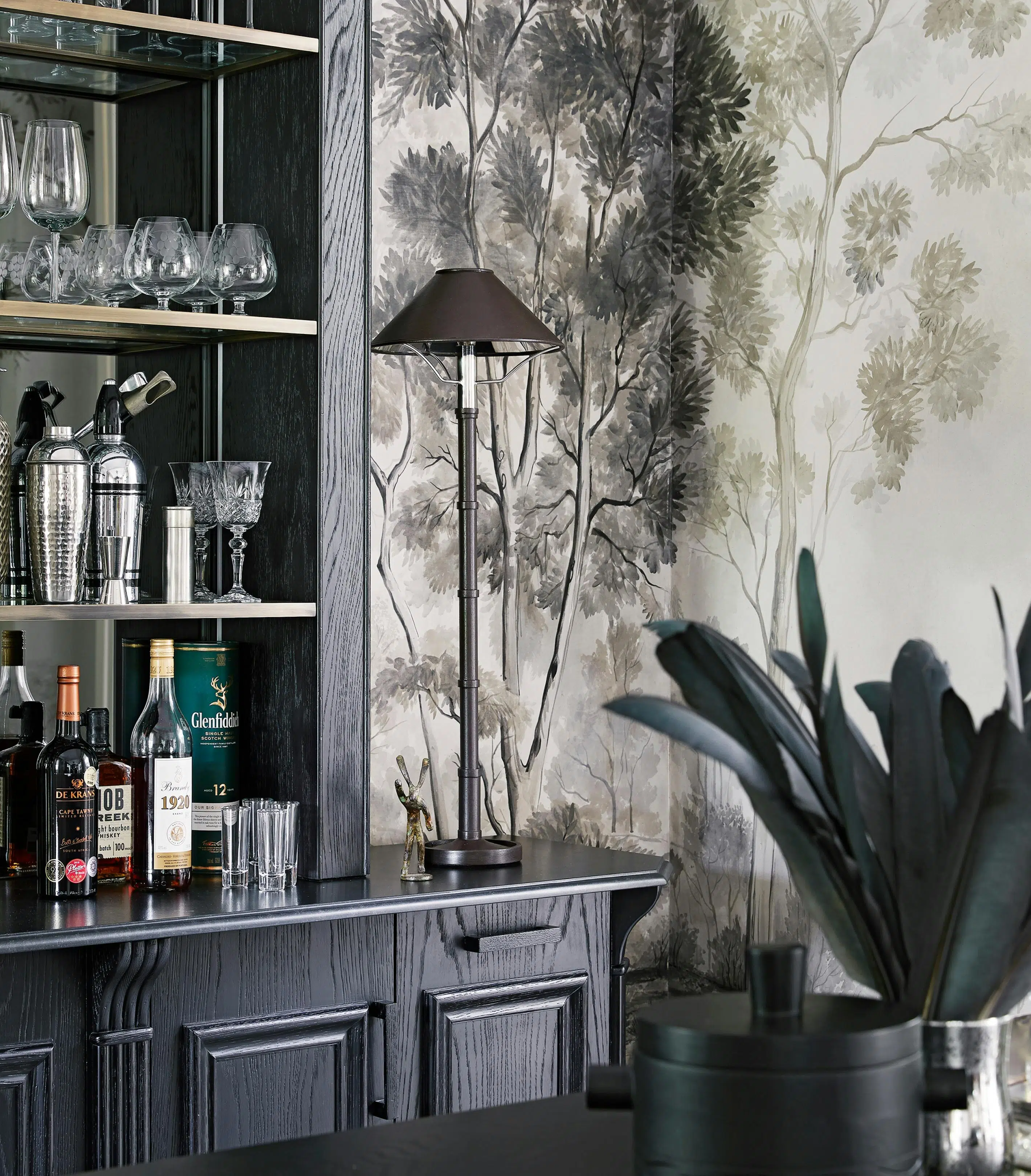 A lamp stands on a tall sideboard holding stemware, cocktail shakers and bottles of whisky, bourbon and brandy. In the foreground is a leafy plant and in the background is wallpaper featuring grayscale watercolour illustrations of trees.