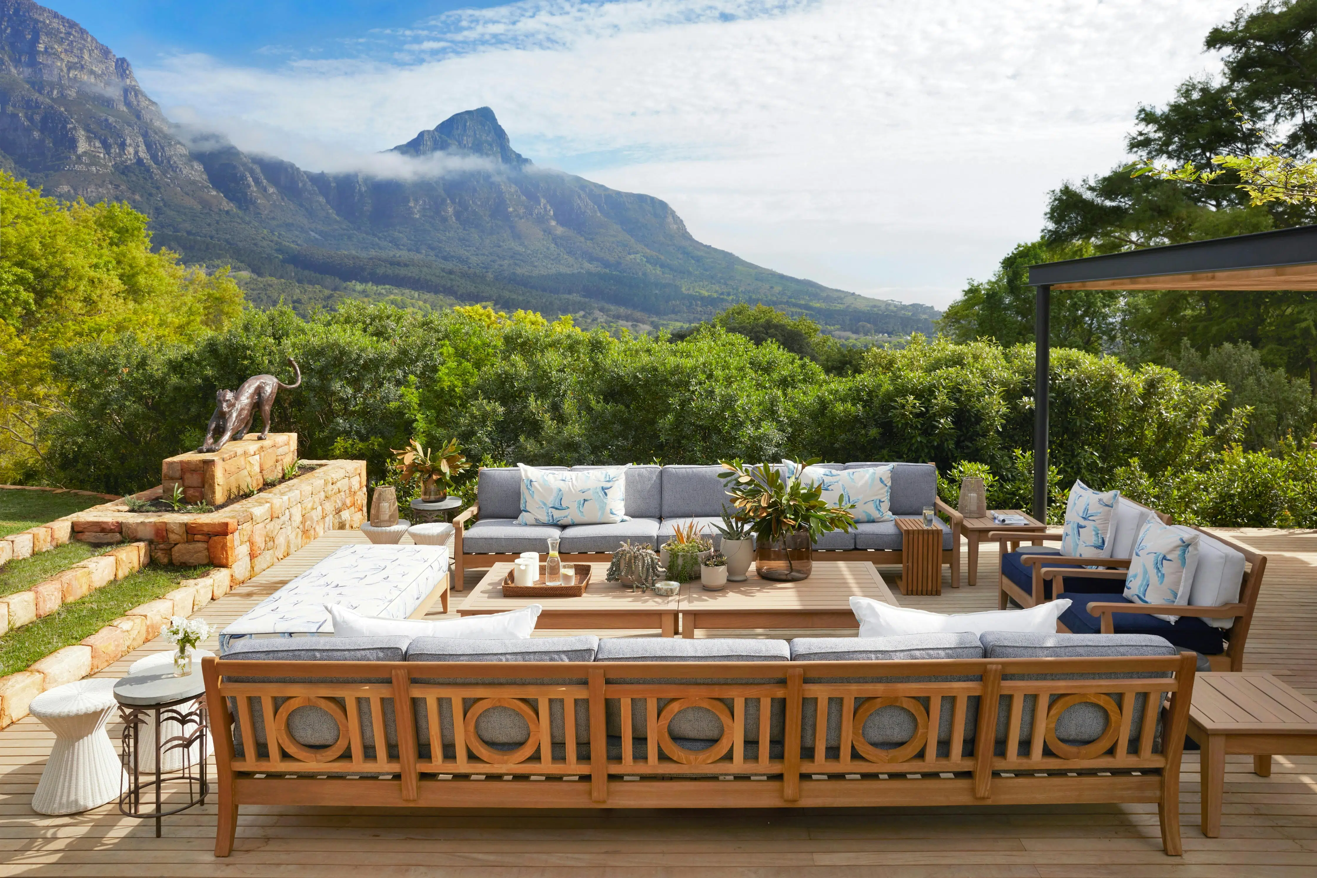 An outdoor seating area with mountain views holds a number of wooden-framed sofas, a central table holding a collection of potted succulents, and a few side tables made of wood, metal and other materials. A metal sculpture of a stretching big cat is visible to the left.