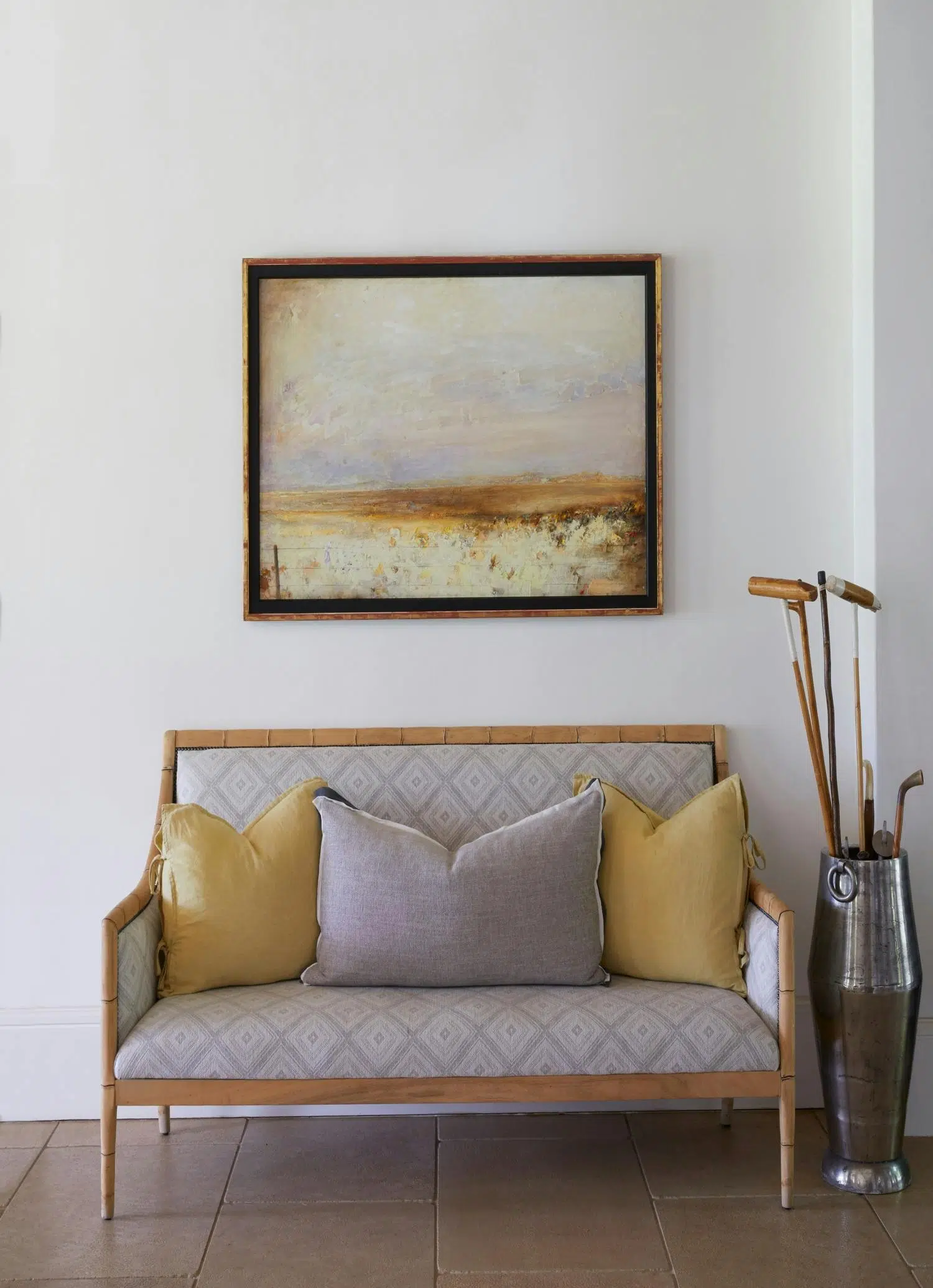 A bench upholstered in grey, geometric-patterned fabric has a light wood frame that seems to be bamboo, and holds three large cushions. It sits on a floor made of large stone tiles, with a metallic vessel beside it holding vintage sporting equipment, and a framed painting above it showing an impressionistic landscape.