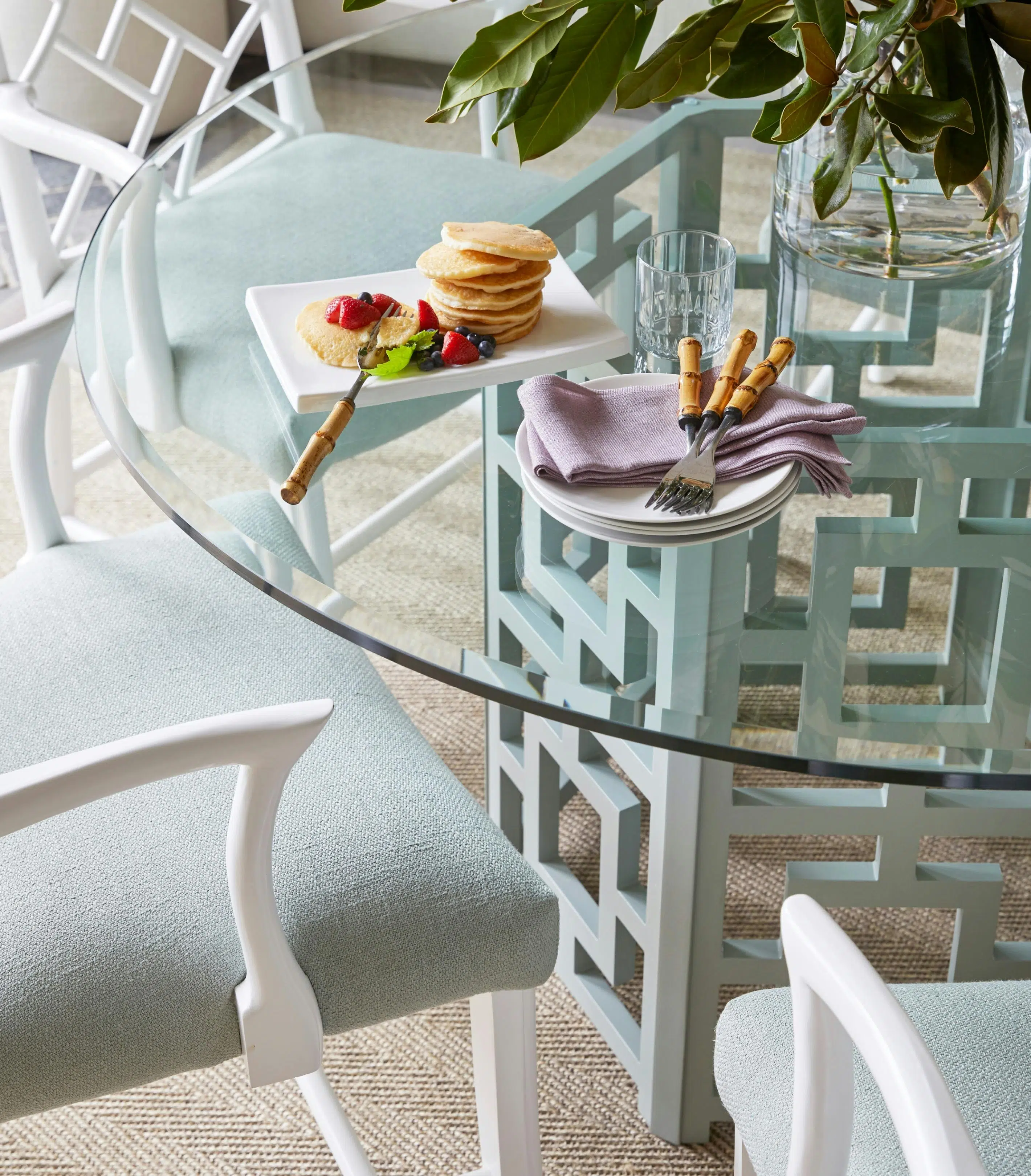 A stack of pancakes and fruit sits on a plate on a round glass table, beside a pile of circular plates holding fabric napkins and forks with handles resembling bamboo. Also on the table are a drinking glass and a glass vase of greenery, while the table's base and accompanying chairs are white, geometric cut-out shapes