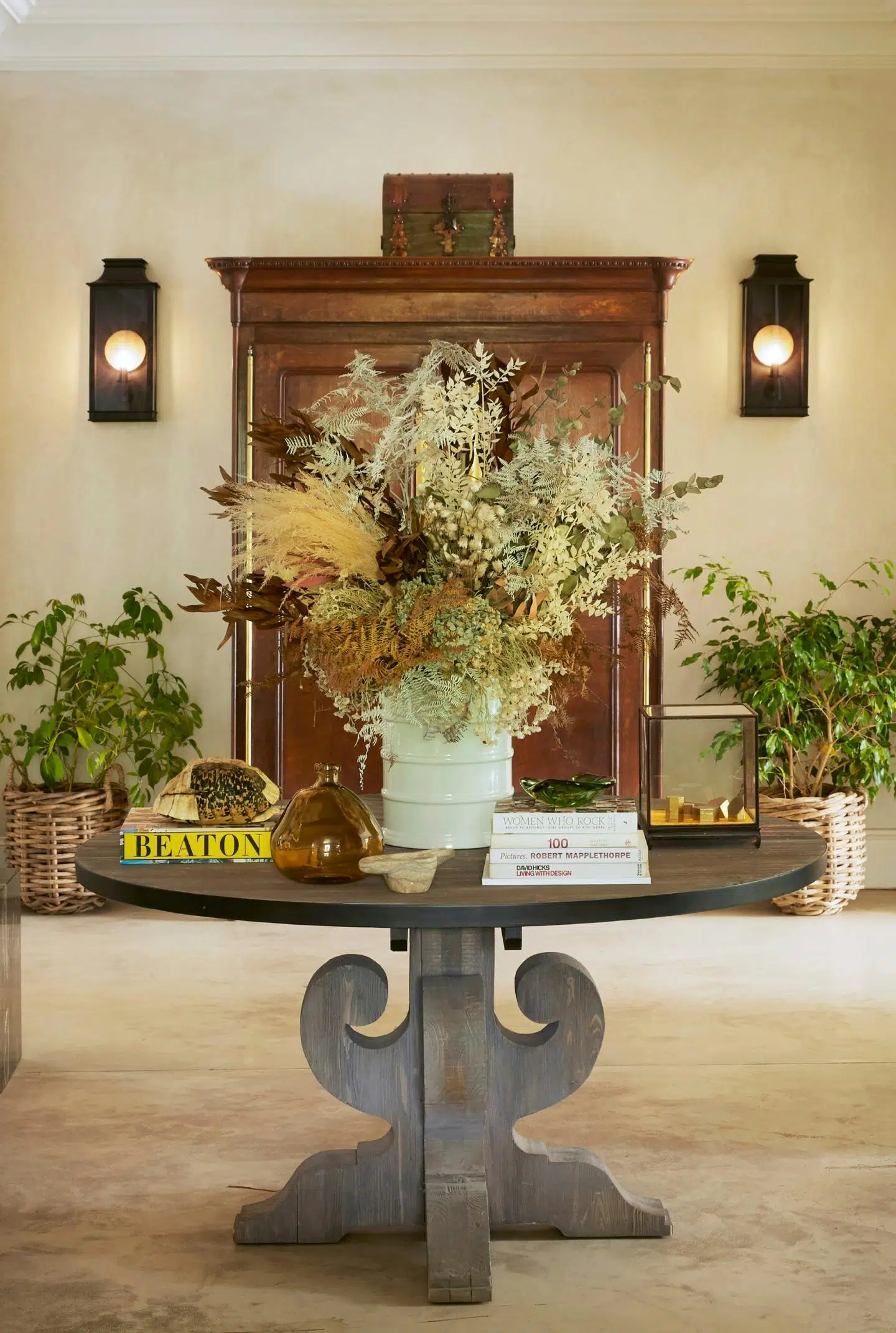 A room is decorated in natural colours and textures with a heavy, round wooden table at its centre, holding art books, glass items and a vase of dried grasses.