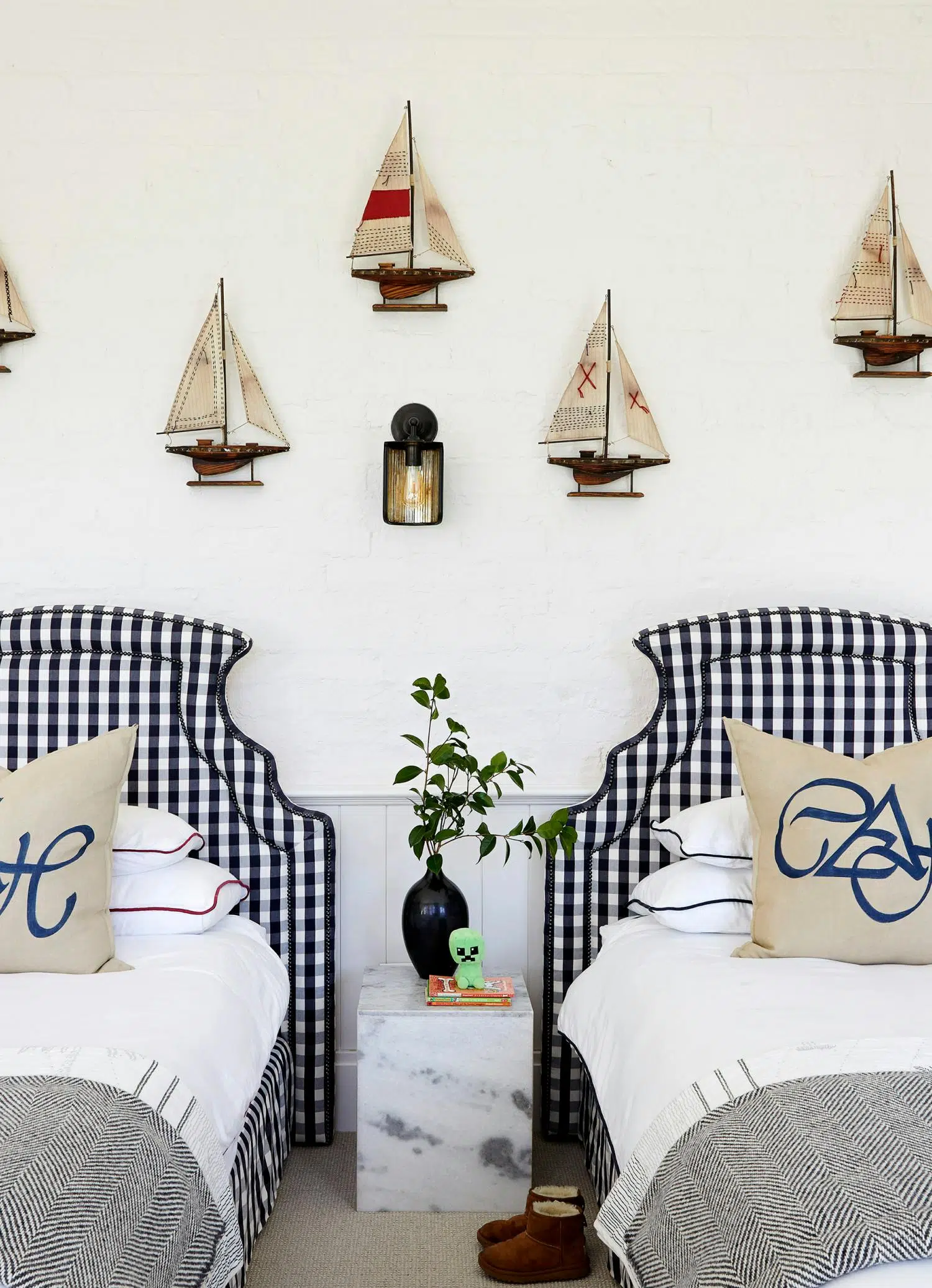 A marine-inspired bedroom with two single beds that both feature blue and white checkered headboards. Artwork of ships hang on the wall with a side table in between the beds.