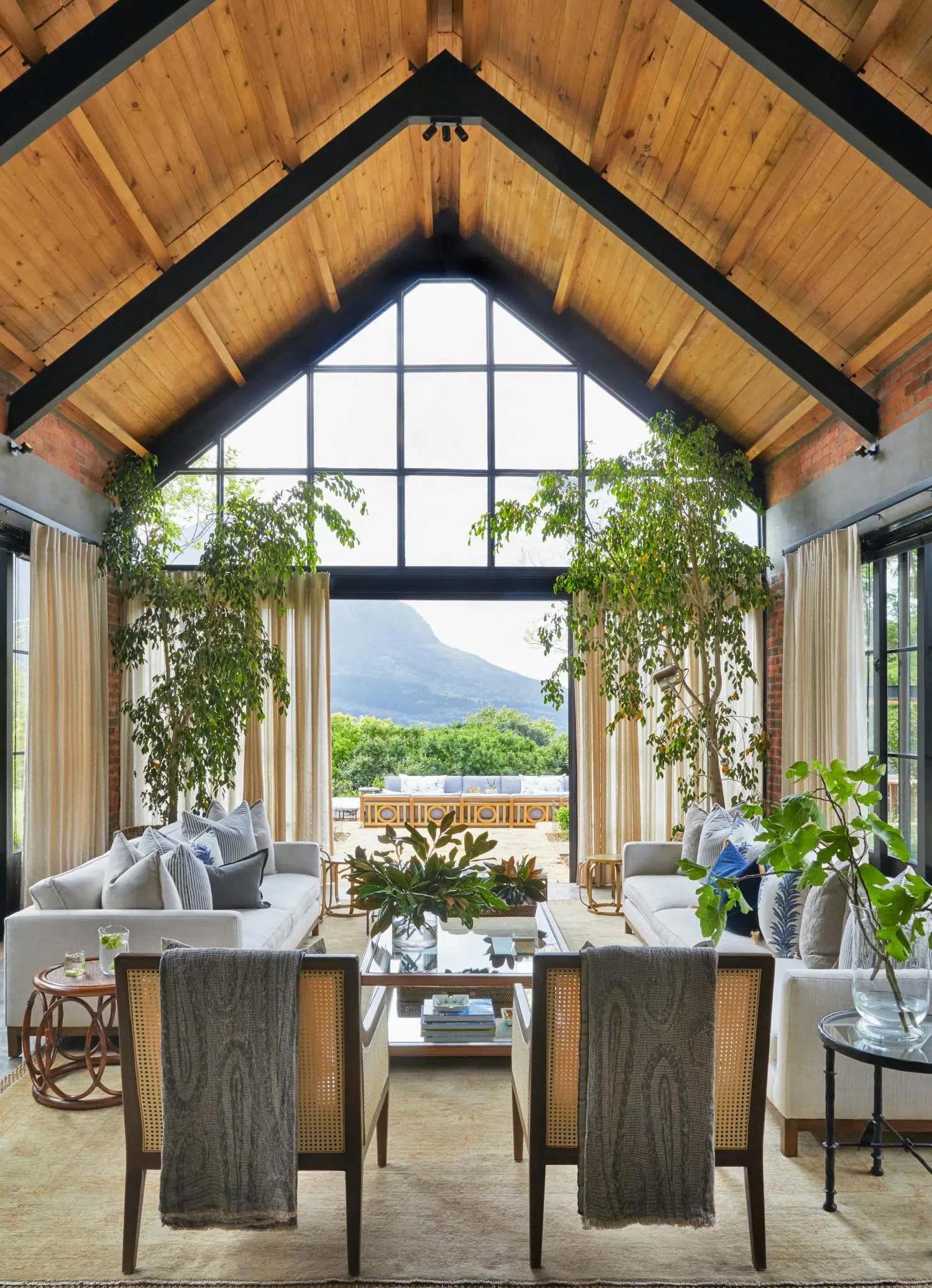 A living room with an A-frame roof, two occasional chairs and two sofas in the foreground with large plants that frame the door to the outside area in the background.