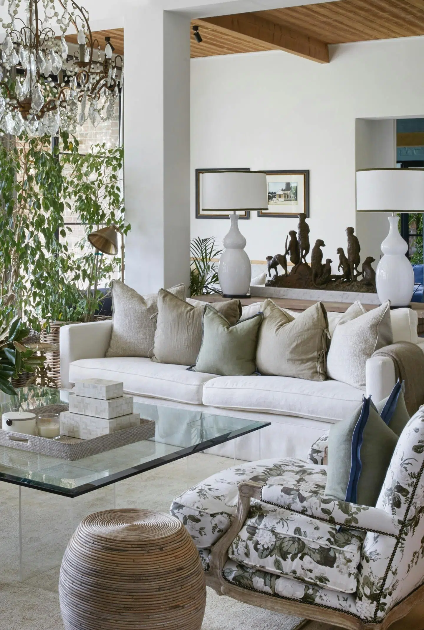 A sitting area features two couches, upholstered chairs, and a glass coffee table holding a tray of square objects and a glass vase of greenery. A leafy plant, a pair of white lamps, and a sculpture of a group of meerkats are visible in the background. A chandelier hangs above