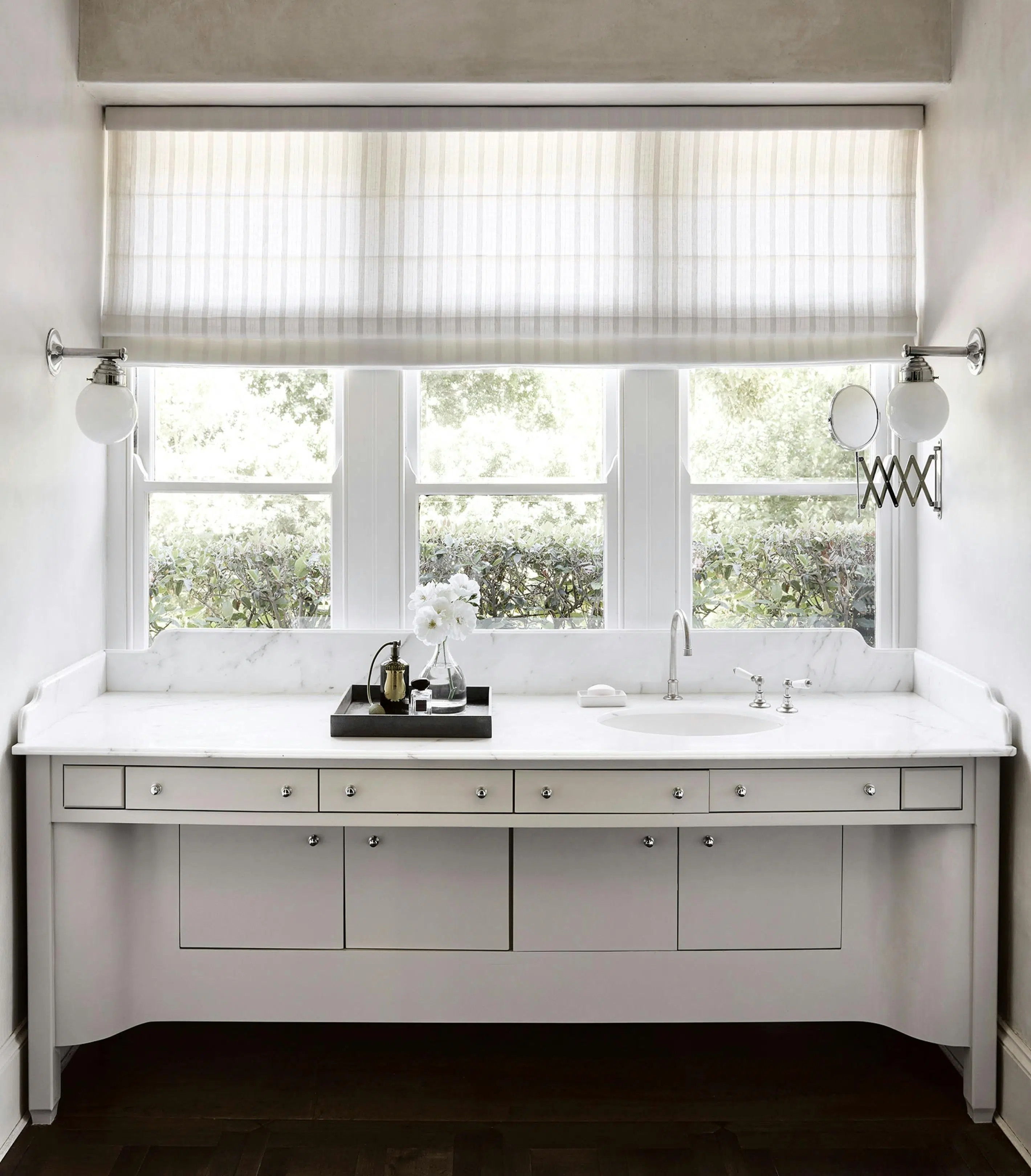 A white bathroom interior looks out through windows onto a garden hedge. A wide vanity's countertop features a sink to the viewer's right, with a tray holding flowers and scent containers to the left.