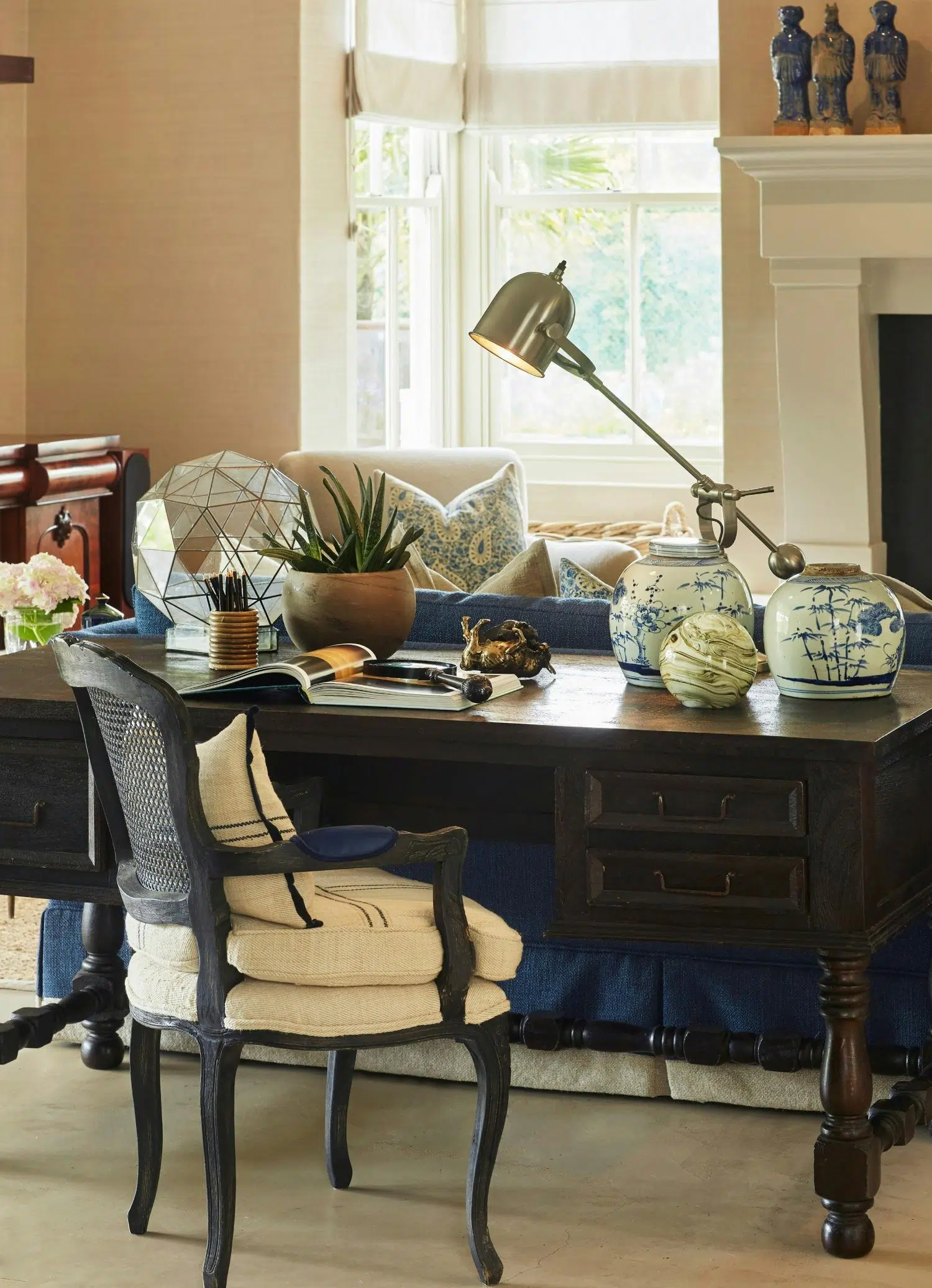 A dark wooden writing desk is in a comfortably furnished room with ornaments, a plant, a desk lamp and an open magazine displayed on its surface. A dark wood Louis XV-style chair in cream fabric stands in front of it, and in the background, three ceramic figures stand on a mantlepiece and a large window sheds light on the room through white blinds.