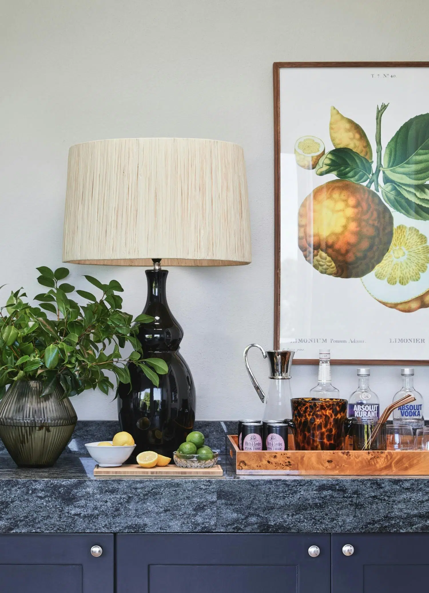 A lamp, a tray of alcohol, a plant is seen on a console table with fruit artwork on the wall just behind.