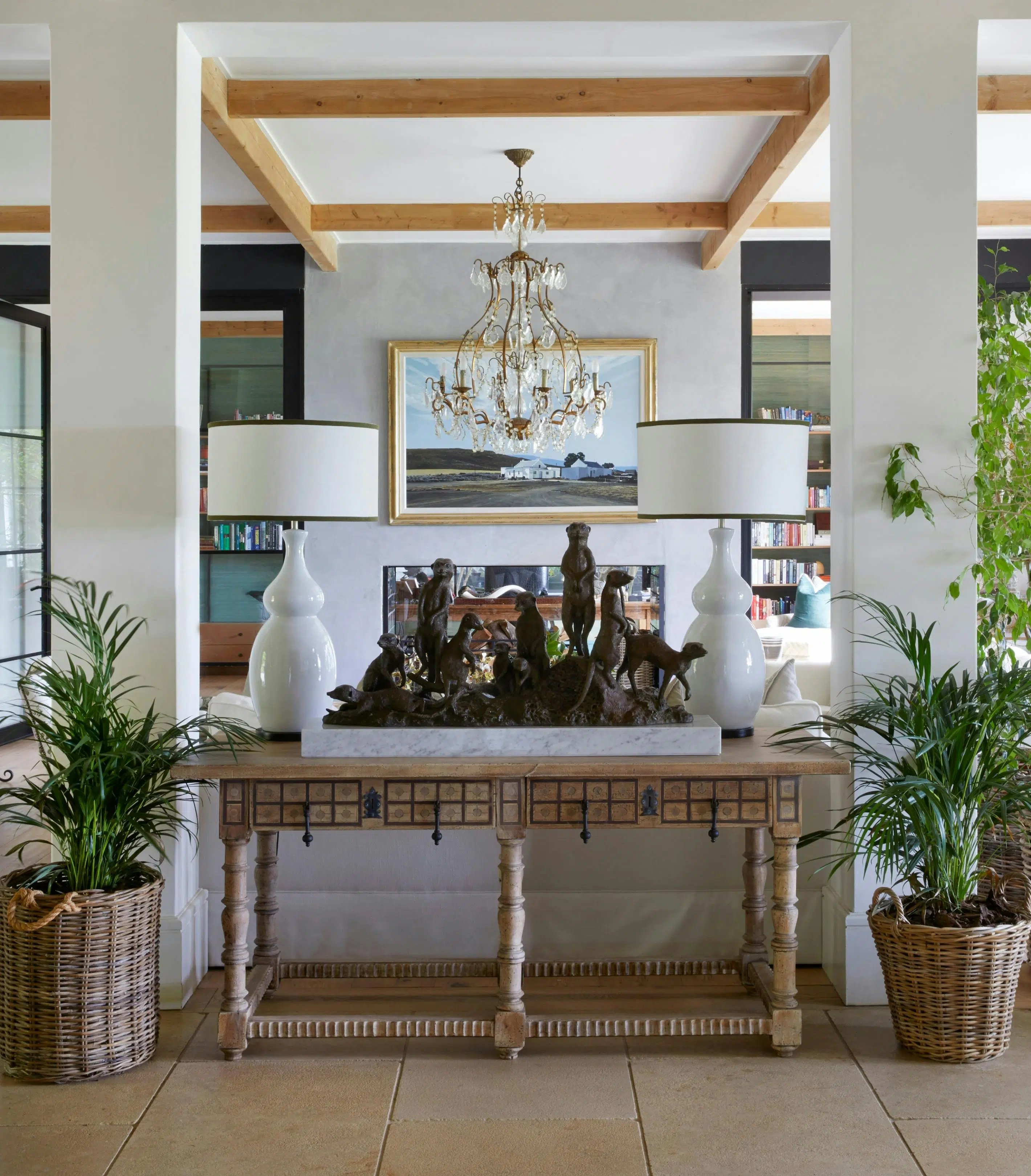 A sculpture of a group of meerkats sits between two large, white ceramic lamps on a wooden console table. A palm in a wicker basket stands on each side of the console, and a chandelier hangs above. A framed painting of a house in a rural setting is visible in the background