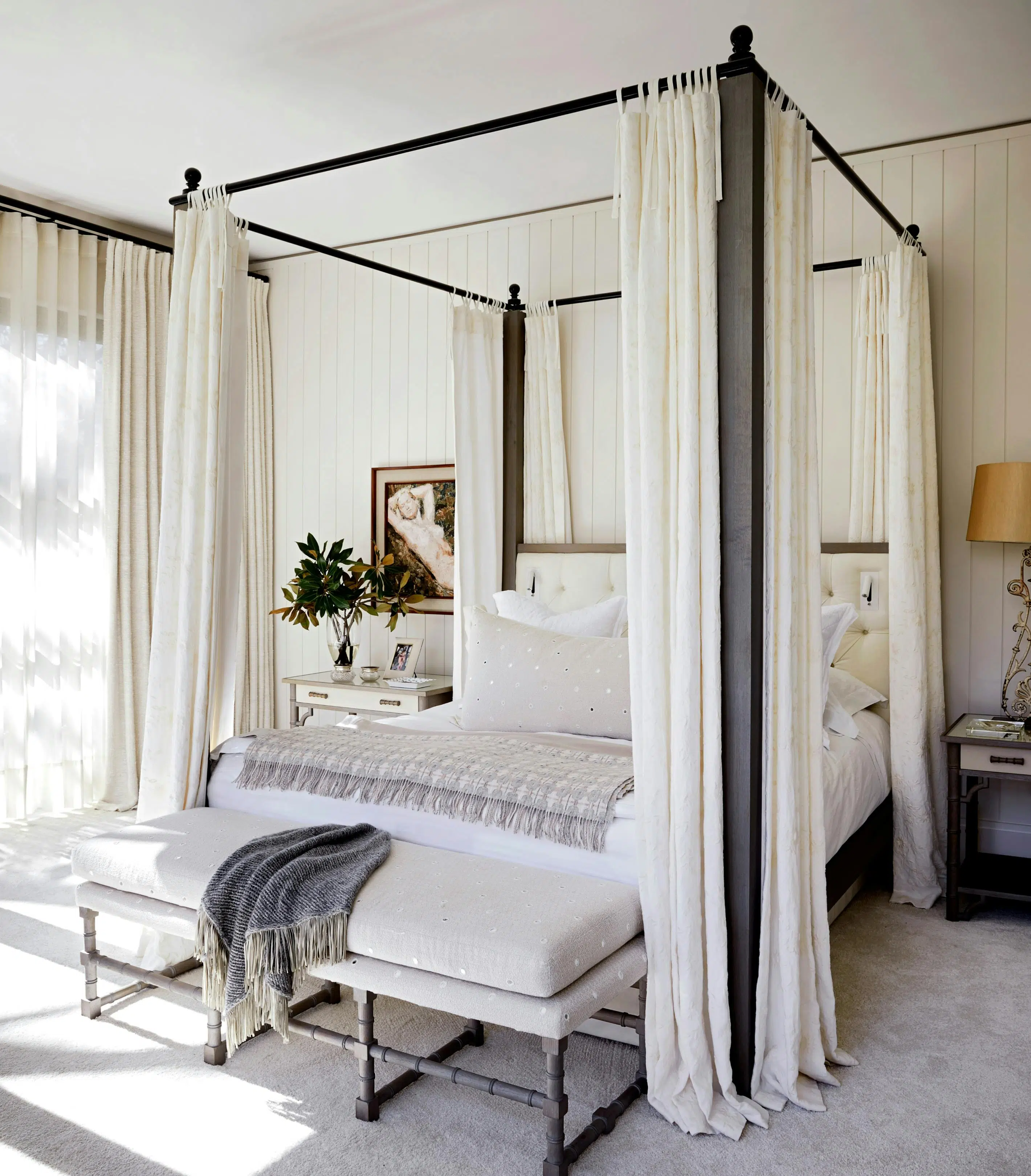 A bedroom that features a sophisticated 4-poster bed in a neutral colour palette of whites and greys with an ottoman just in front of the bed.