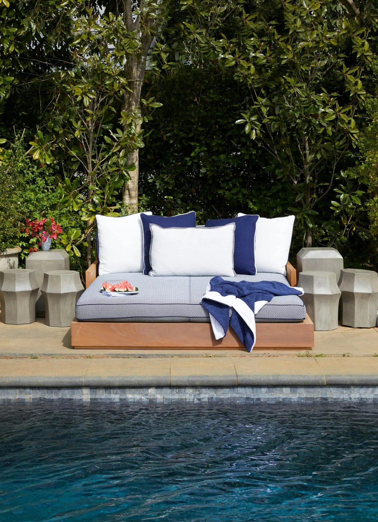The water of a dark-bottomed swimming pool is visible in front of poolside furniture holding blue and white cushions, a blue-and-white towel, and a plate of watermelon slices. Lush greenery is in the background.