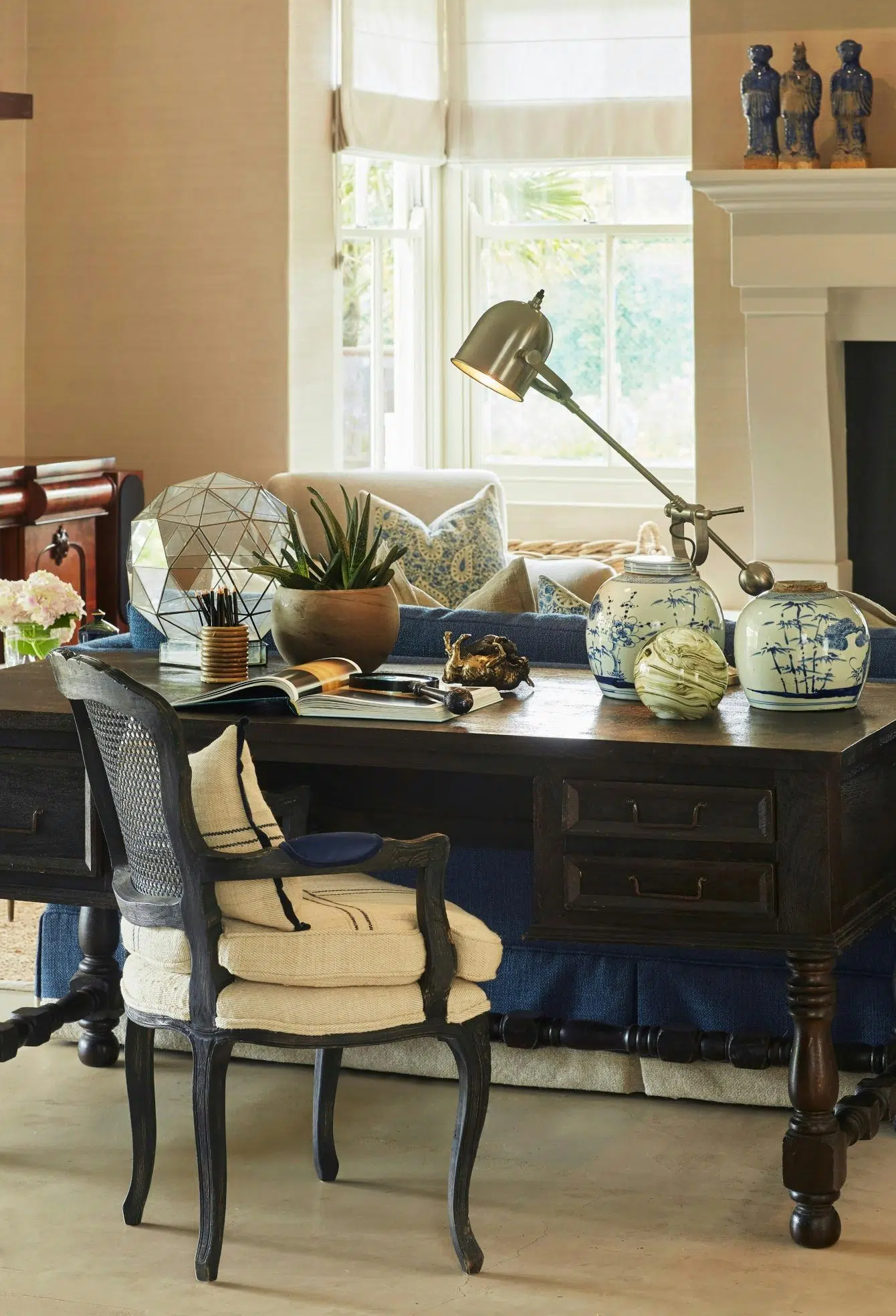 A dark wooden writing desk is in a comfortably furnished room with ornaments, a plant, a desk lamp and an open magazine displayed on its surface. A dark wood Louis XV-style chair in cream fabric stands in front of it, and in the background, three ceramic figures stand on a mantlepiece and a large window sheds light on the room through white blinds.