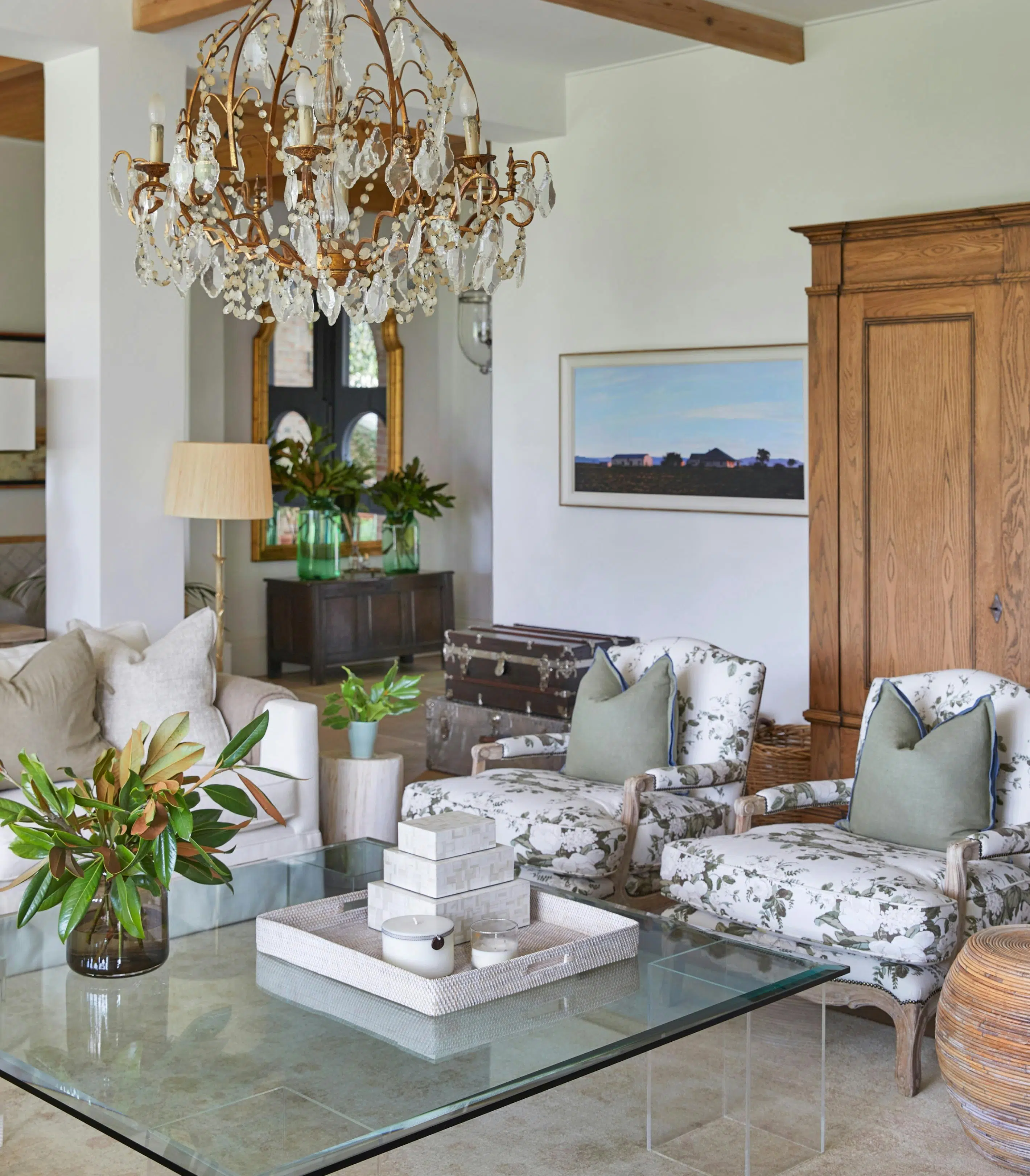 A sitting area features a couch, two patterned, upholstered chairs, and a glass coffee table holding a tray of square objects and a glass vase of greenery. A large wooden wardrobe and a framed painting of a rural town are seen in the background. A chandelier hangs above