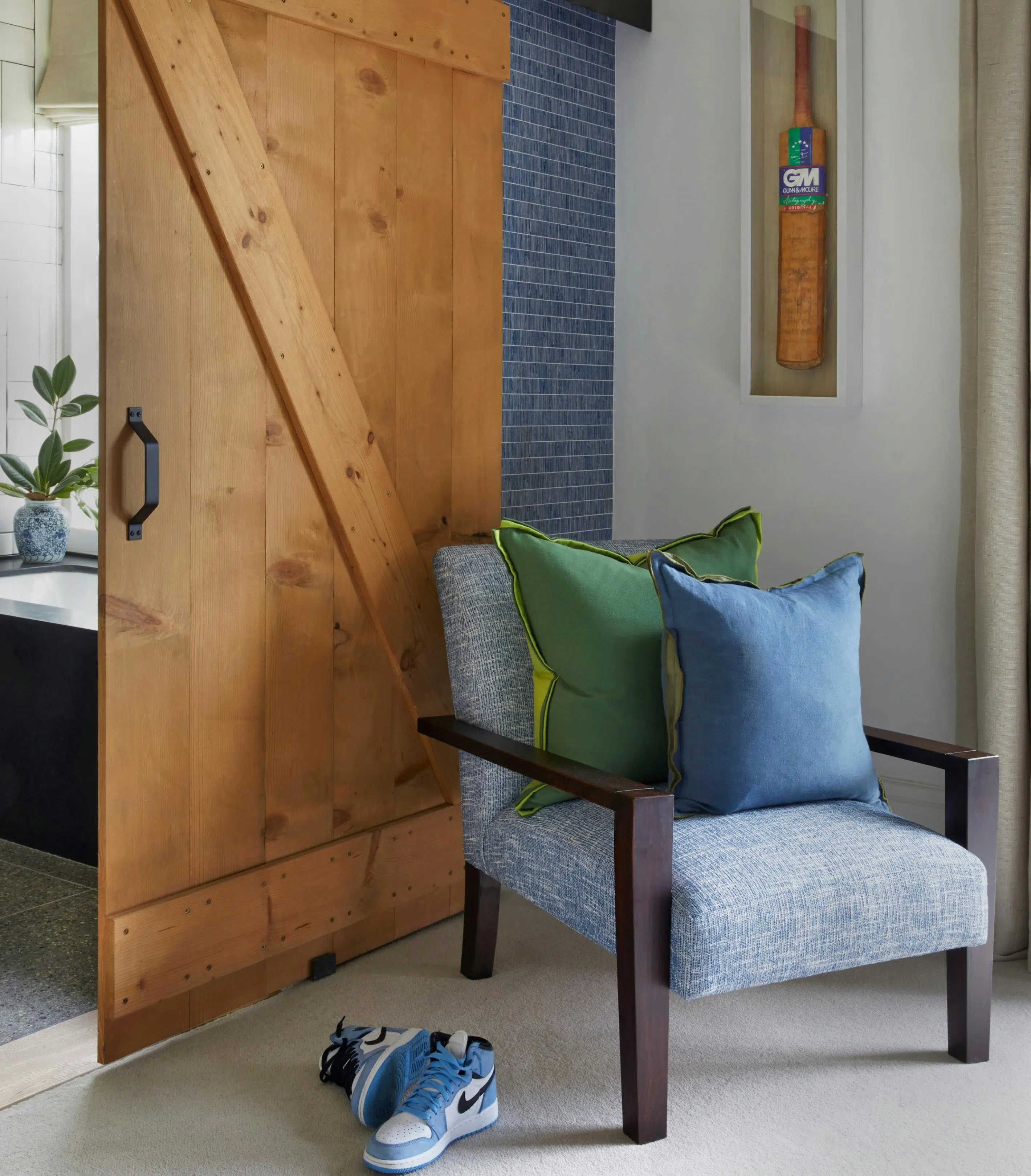 A blue-upholstered armchair with wooden armrests and two cushions – one green and one blue – sits in a corner under a framed cricket bat. A wooden door next to the chair seems to lead to a bathroom, and blue-and-white Nike sneakers are on the floor