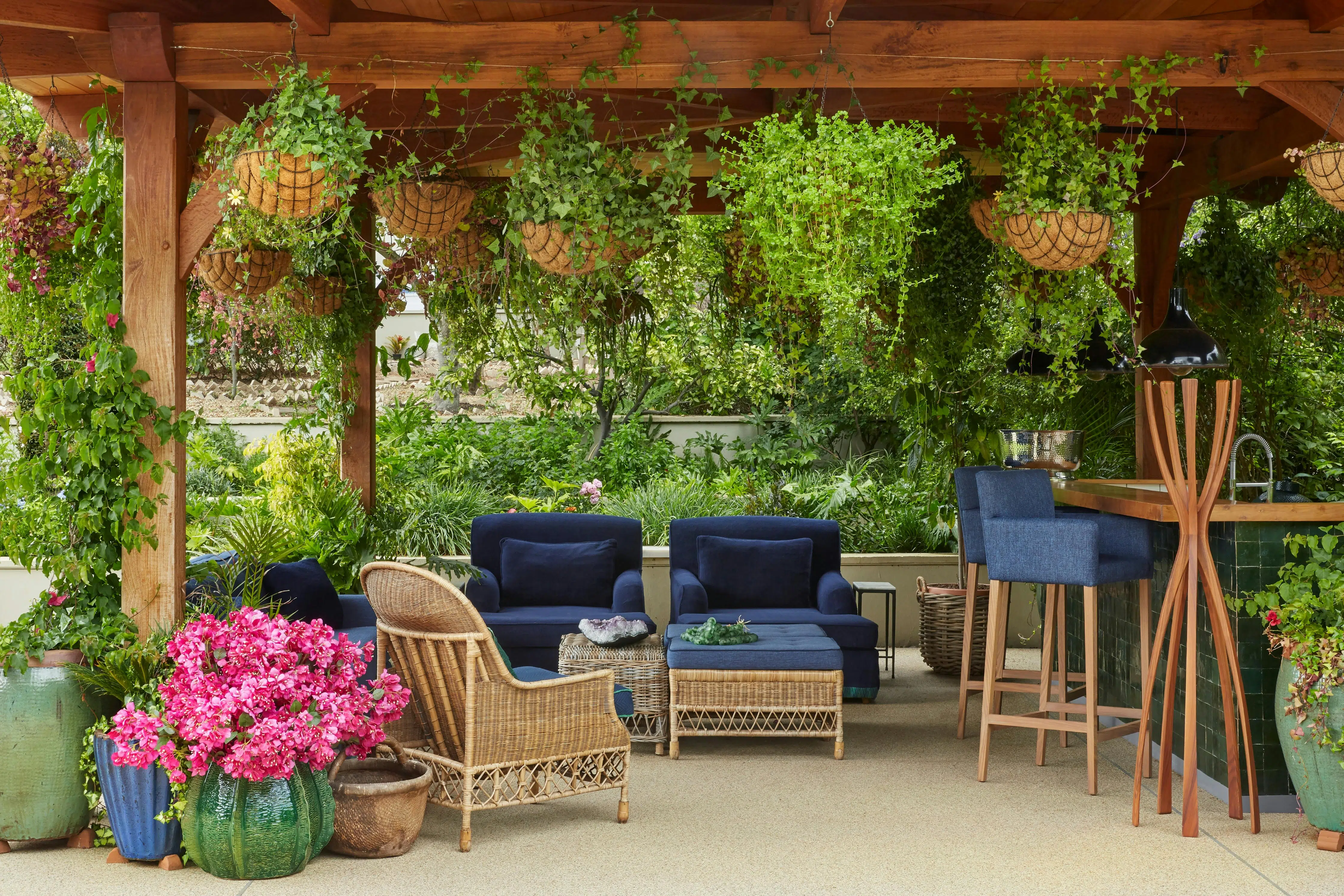 An outdoor entertaining area featuring rattan furniture, surrounded by greenery, with a bar area to the left.