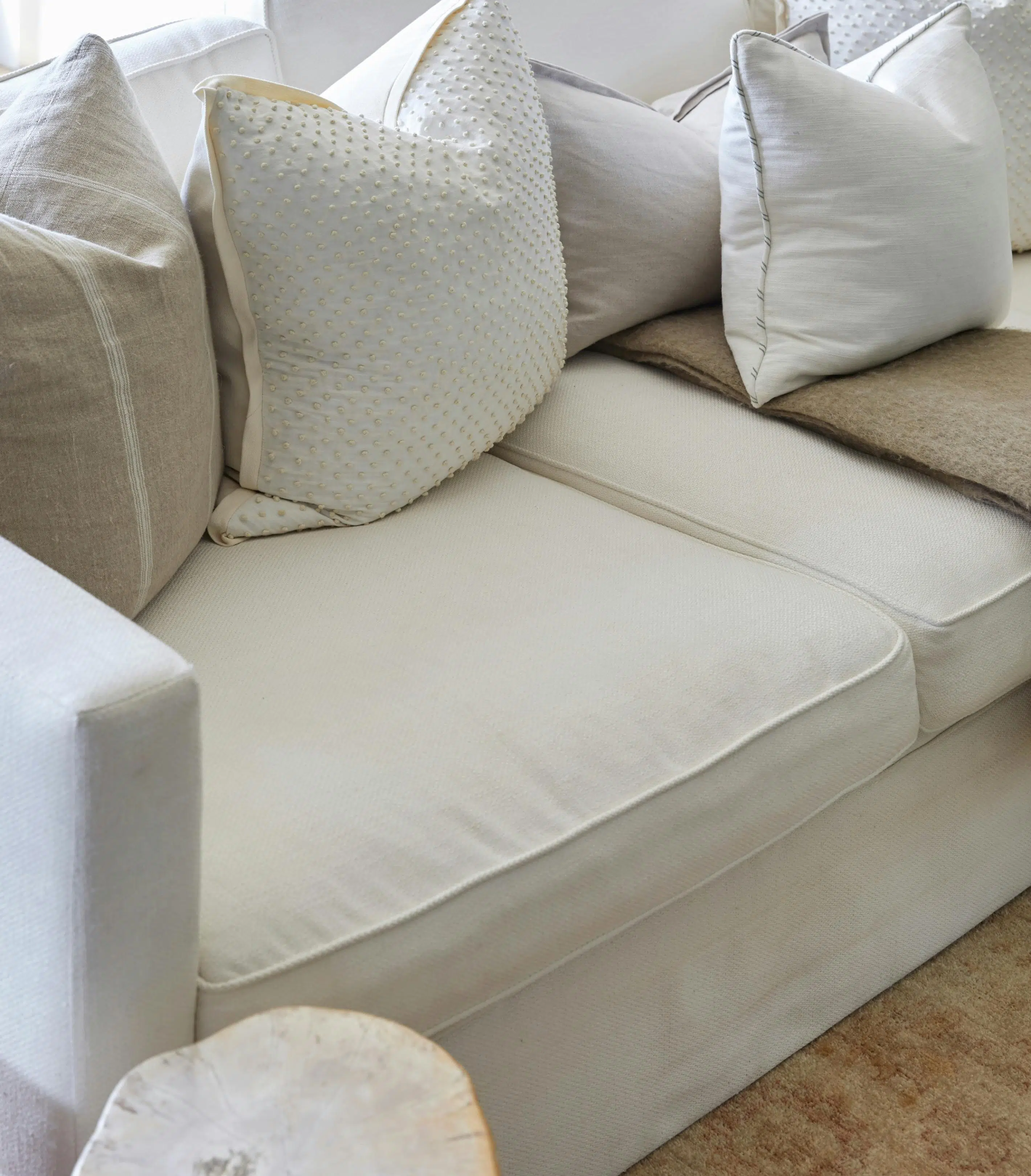 A closeup of a couch upholstered in pale fabric holding cushions in various textures and neutral tones, as well as a folded, light brown blanket