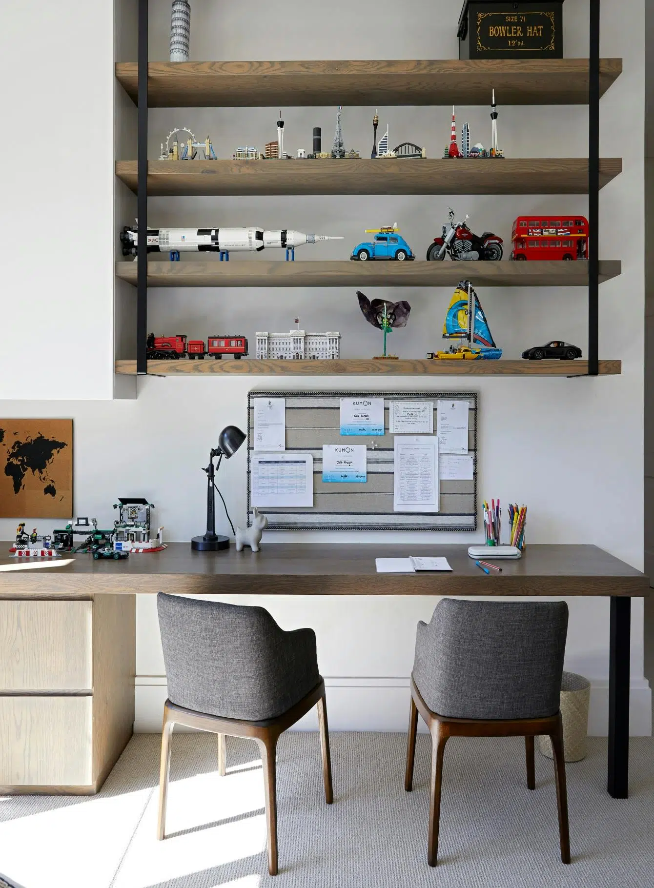 An at-home office that features a wooden desk with two chairs upholstered in grey fabric in the foreground. There is shelving above the desk with decorative accents.