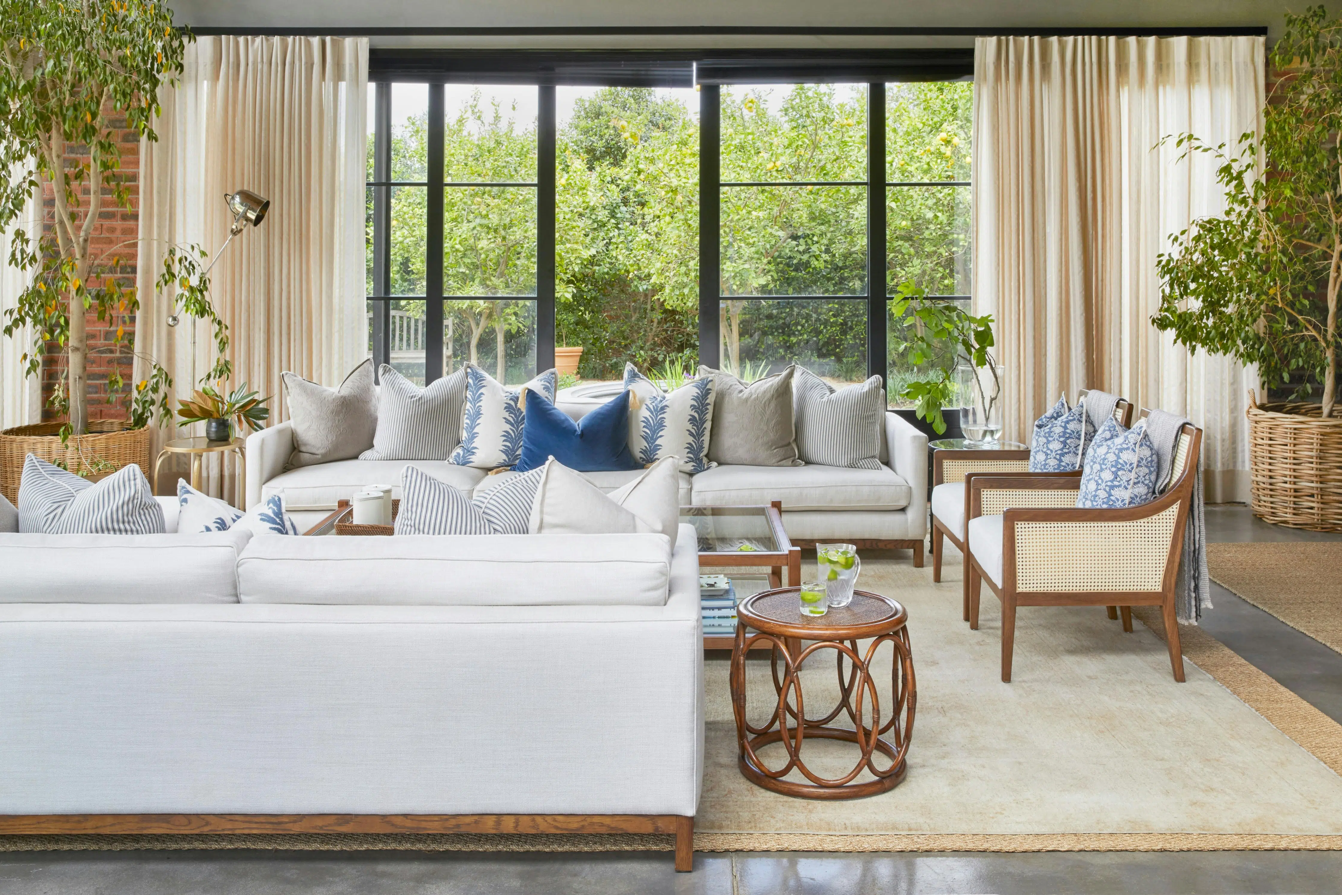 A living area decorated in neutral tones, natural wood, blue-patterned cushions, and fresh greenery. The garden is visible through glass doors