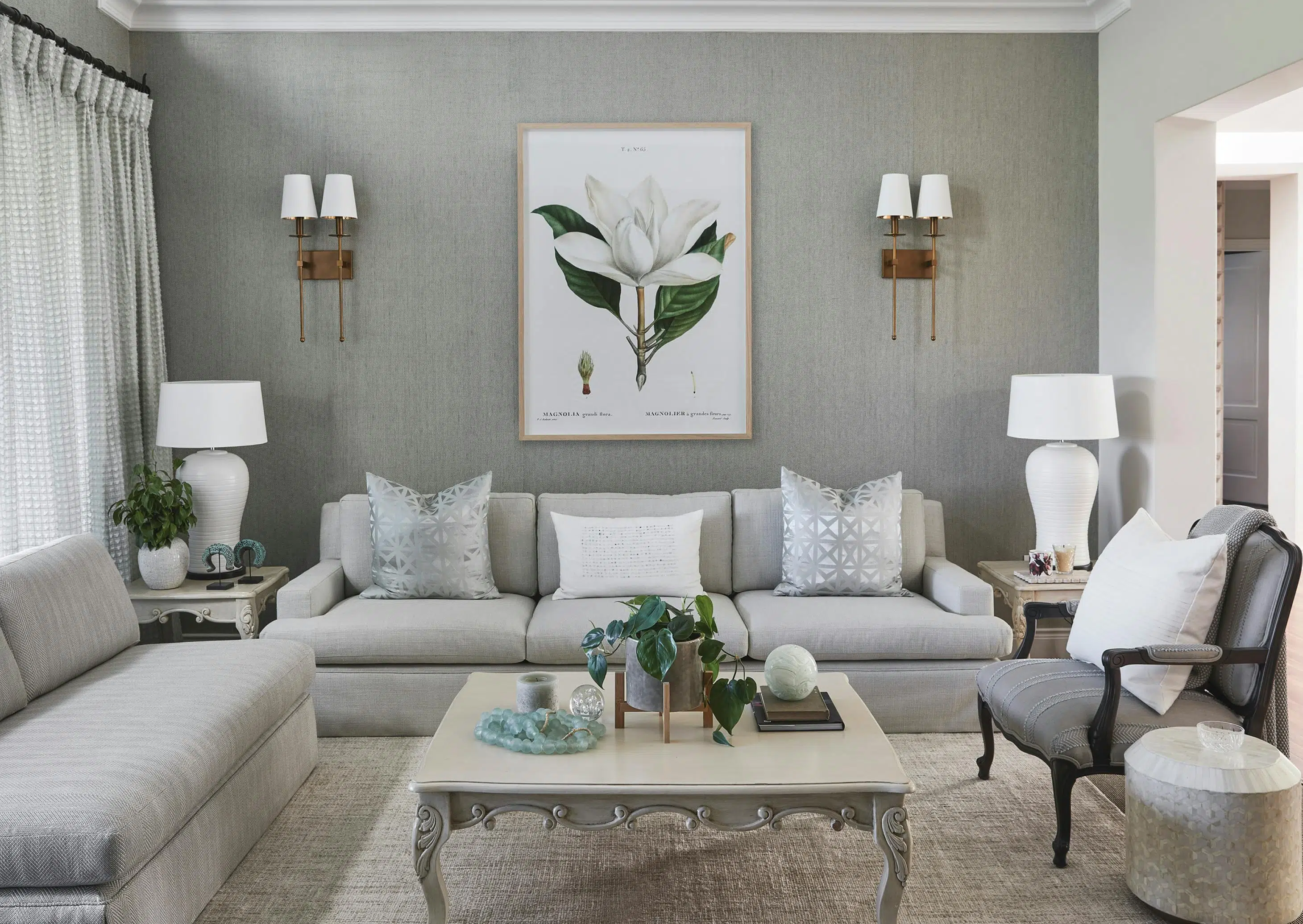 An elegant living room with a neutral, grey colour palette, an ornate coffee table in the middle, surrounded by grey sofas and an armchair with flower artwork on the wall at the back.