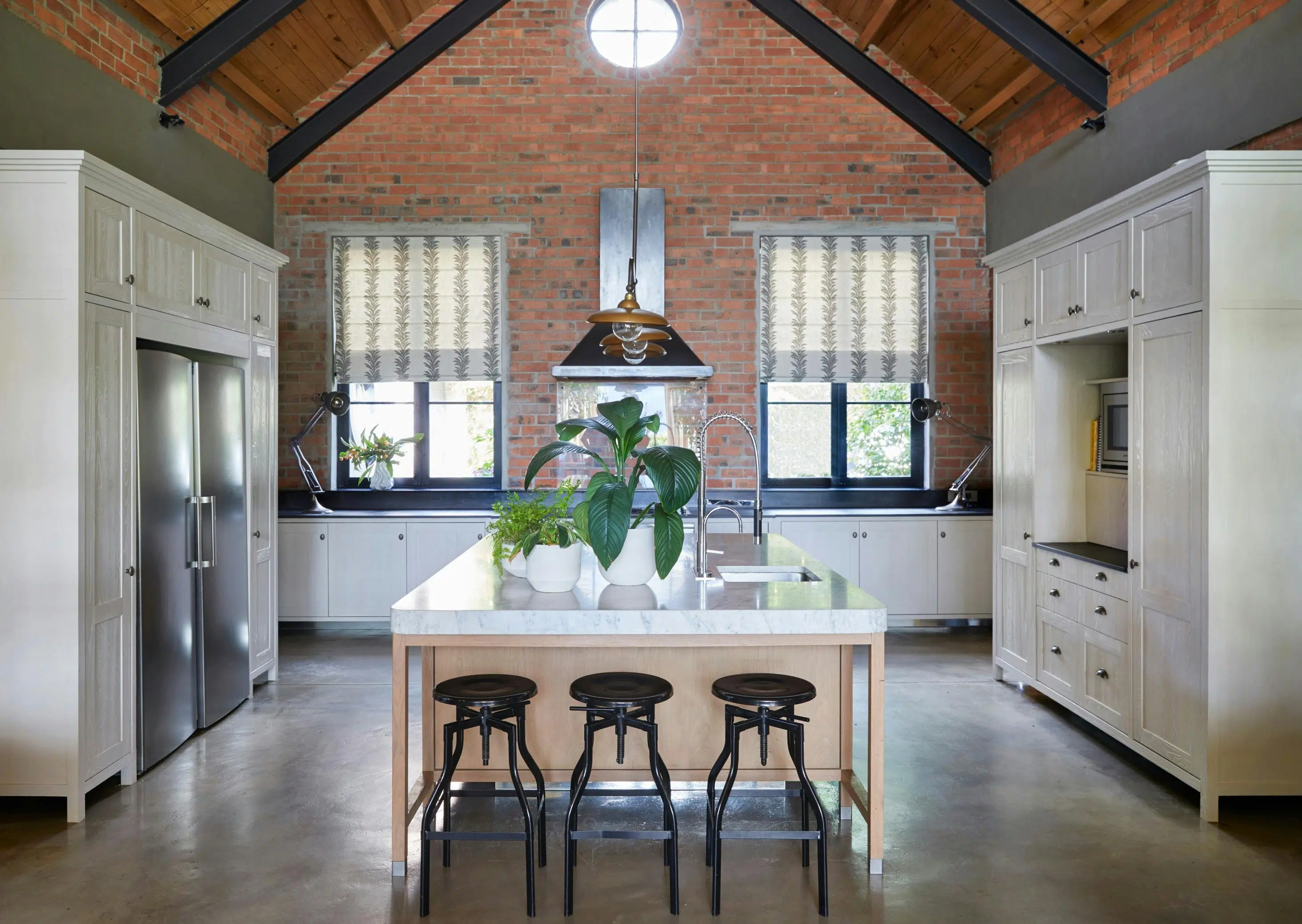 A modern, A-frame kitchen with an exposed brick wall features a central kitchen island and white cabinetry. Bar stools are arranged in front of the island and potted plants sit on its surface.