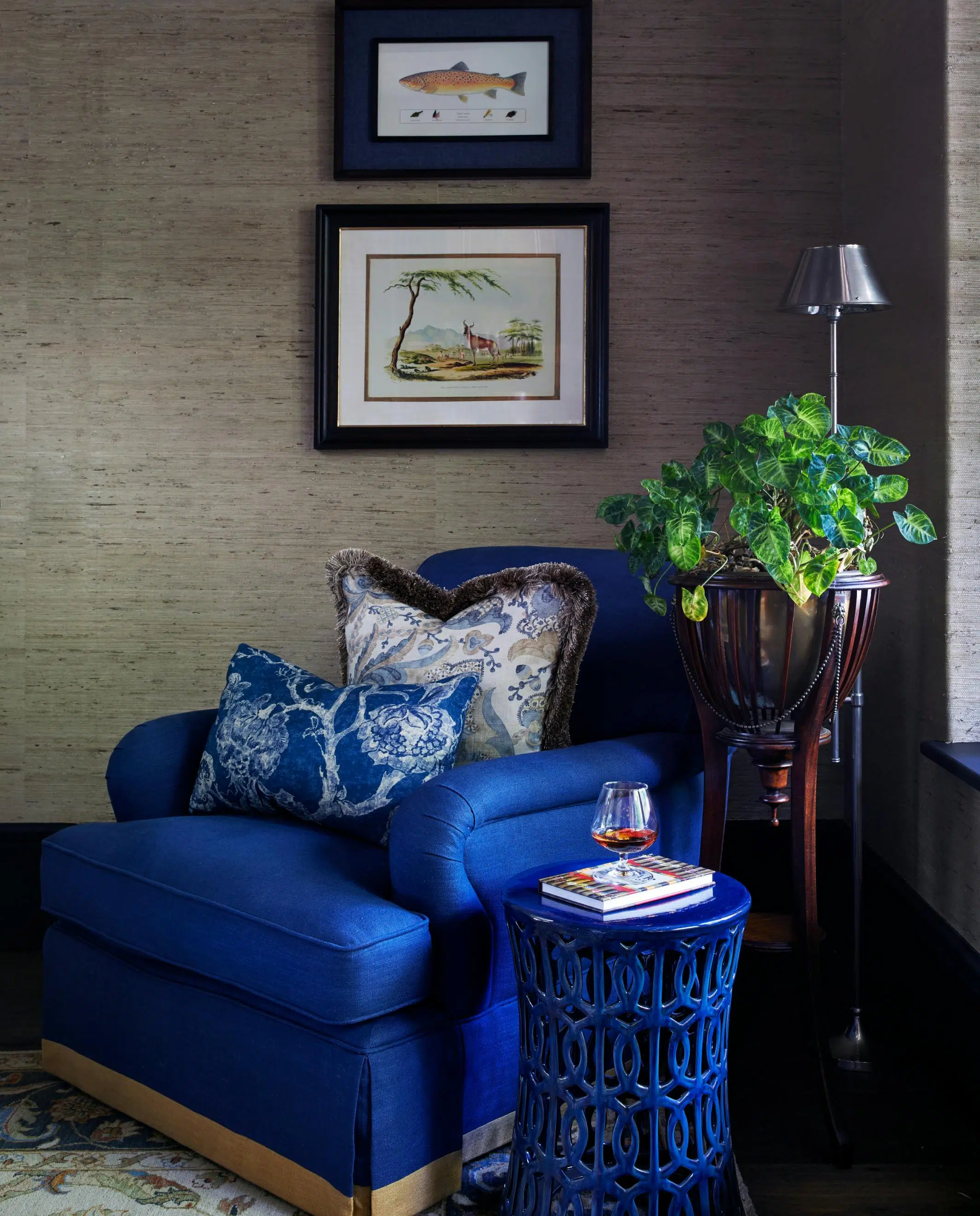 A blue side table with a book and a glass of wine placed on top in front of a blue armchair with marine-inspired artwork hanging above.