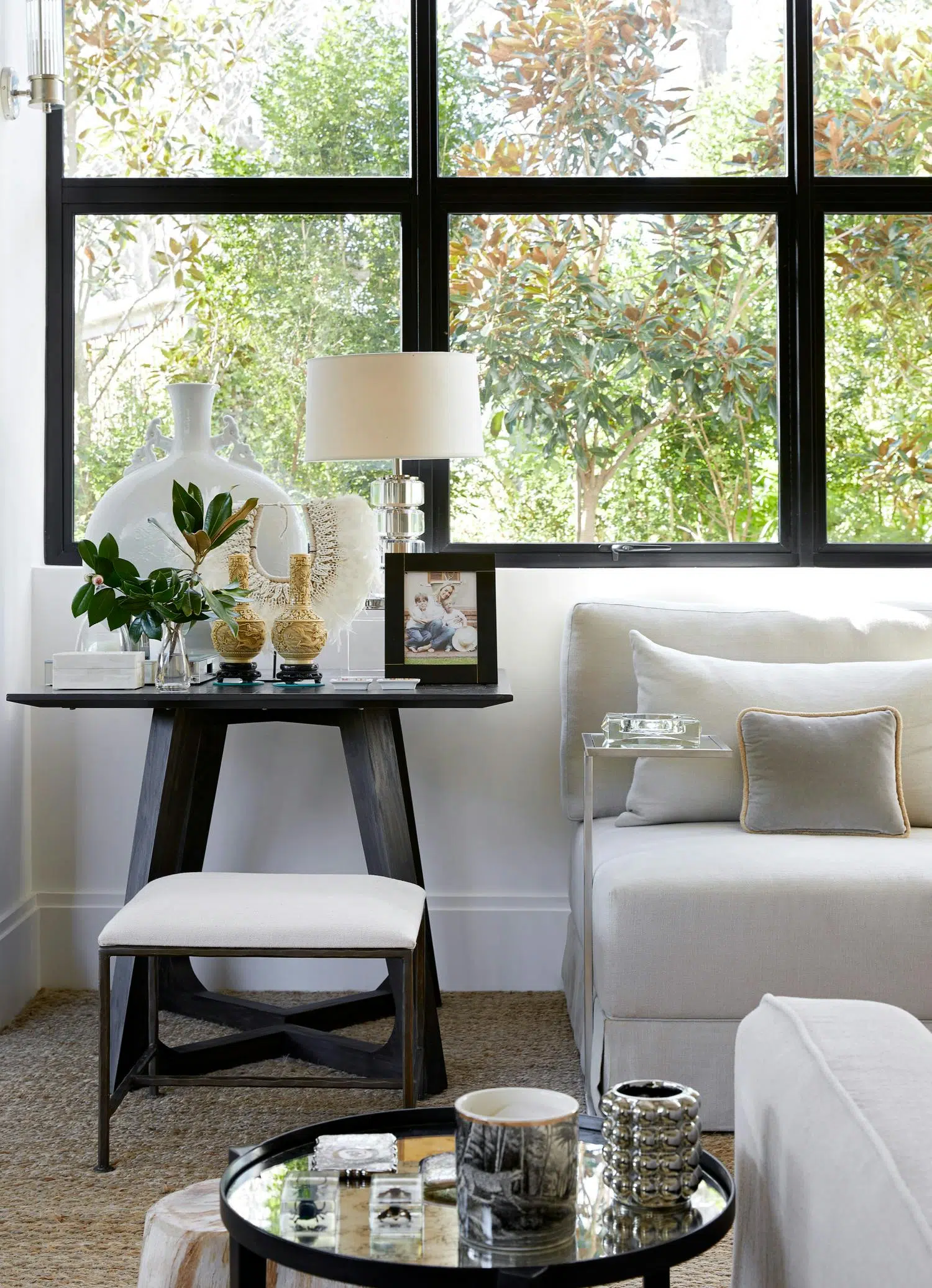 Close-up of a living room with two side tables with decorative accents on top, a window with a view of a tree outside, and the corner of a white sofa.