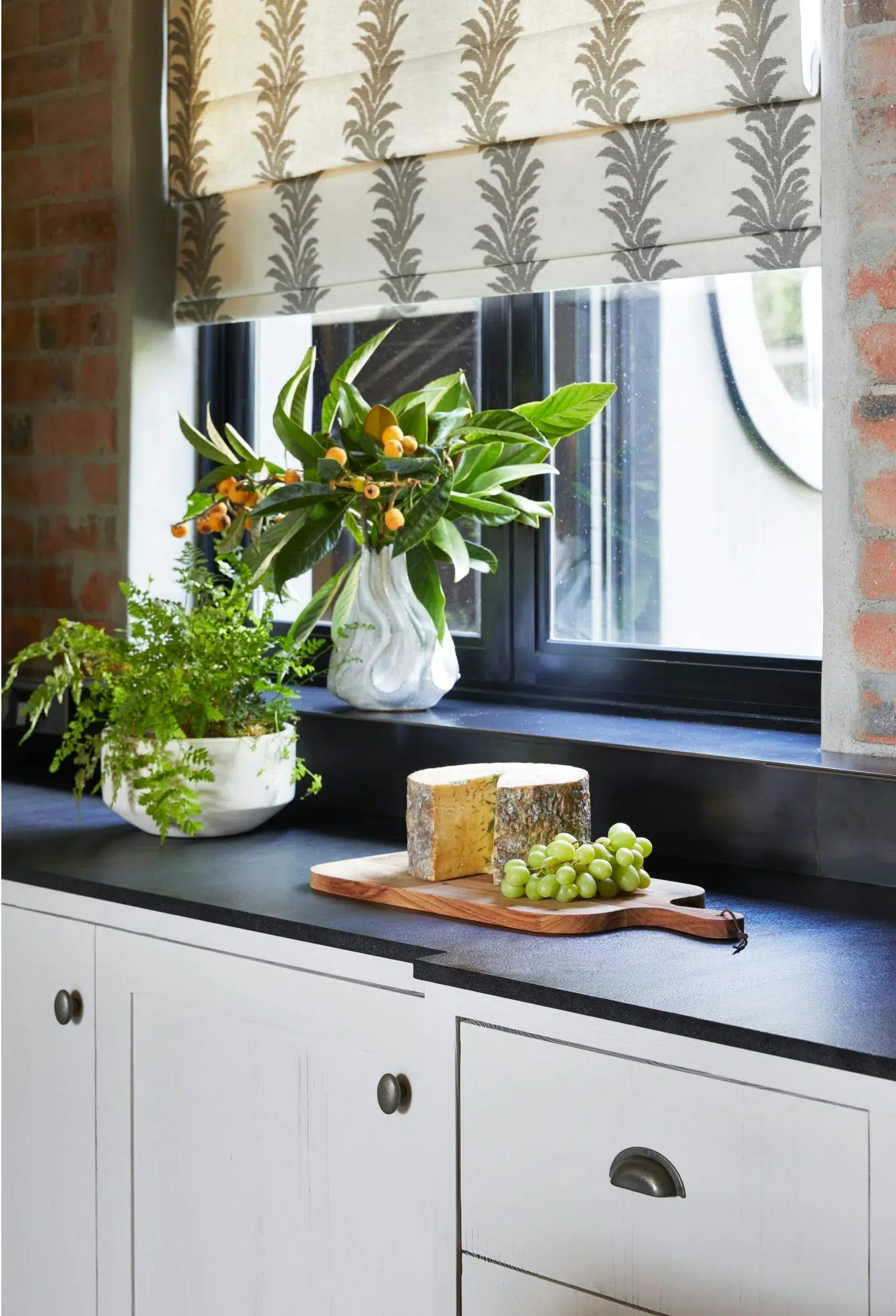 A black kitchen countertop holds a wooden board piled with green grapes and a large wheel of cheese. Behind this is a window draped with blinds in a stylised, botanical pattens, and on the counter and windowsill are a white planter holding greenery and a white organic-shaped vase holding loquat fruit and leaves
