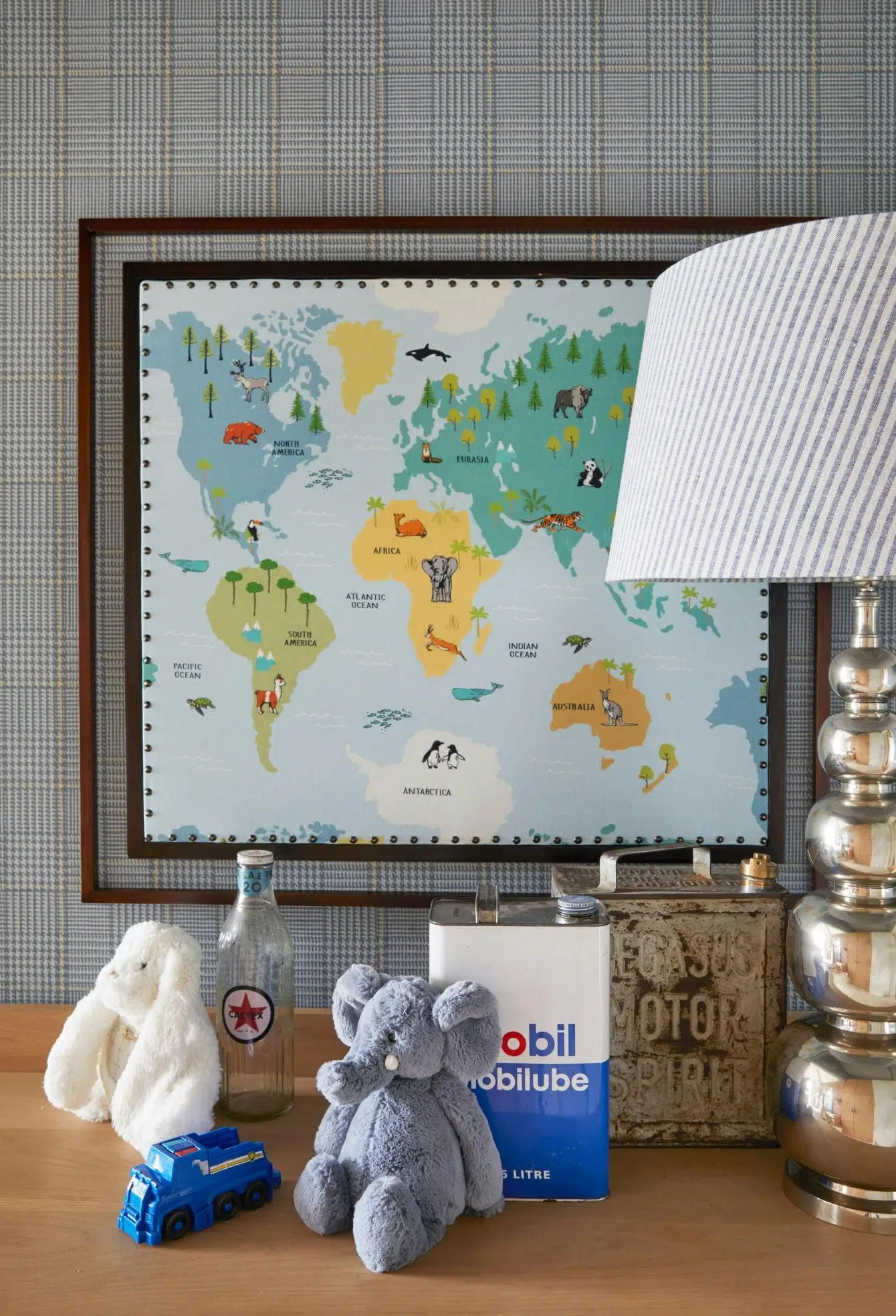 In a closeup from a child's bedroom, a plush rabbit and elephant sit next to two vintage oil cannisters and a small toy truck. A world map illustrated with animals on their native continents is framed on the wall against plaid wallpaper, and a striped lampshade sits on a metallic base.