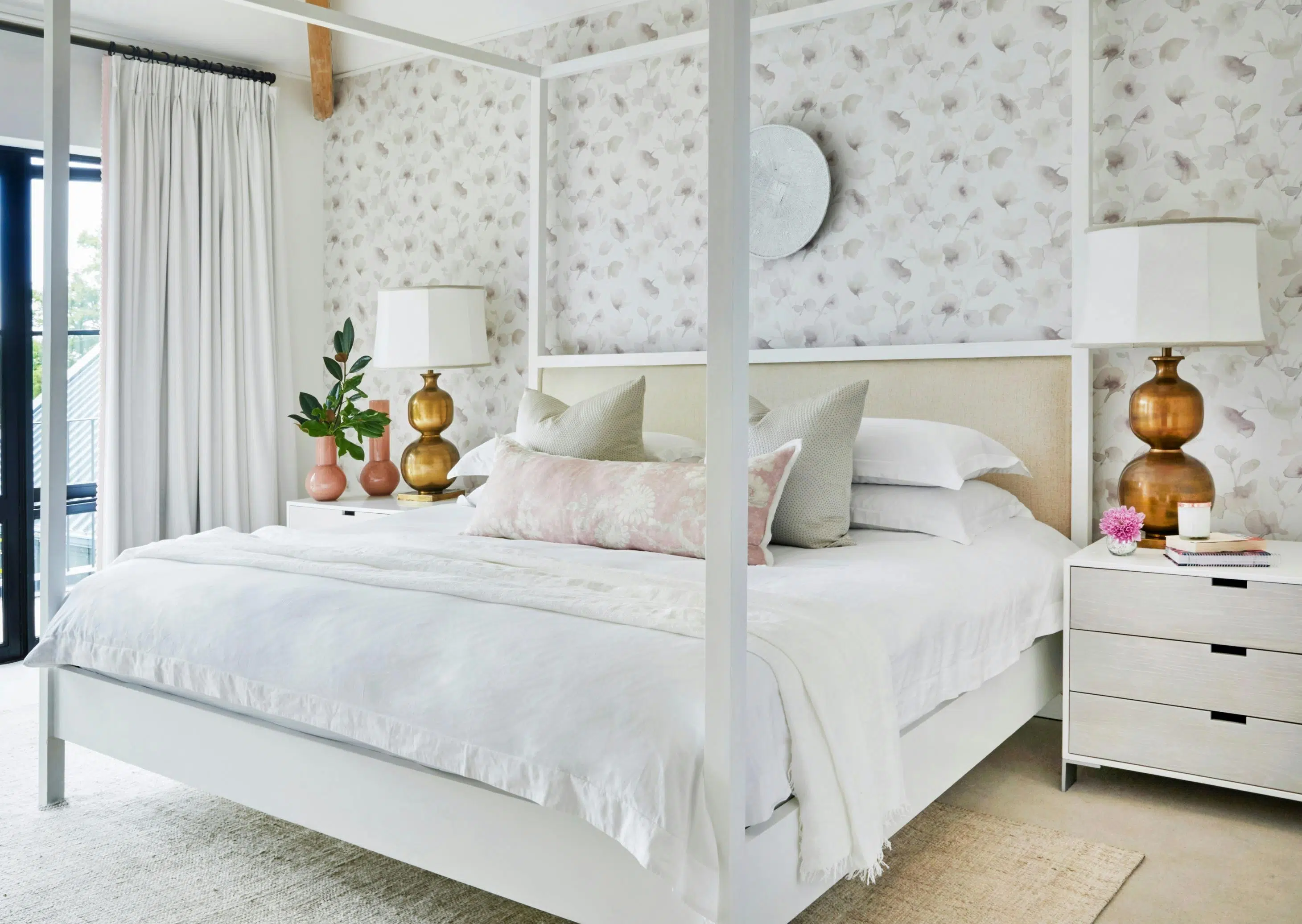 A wide four-poster bed with a white frame, white linen and pale-coloured pillows sits in front of a wall papered in a pale, sepia botanical print. White side tables to the left and right of the bed hold metallic lamps and vases.