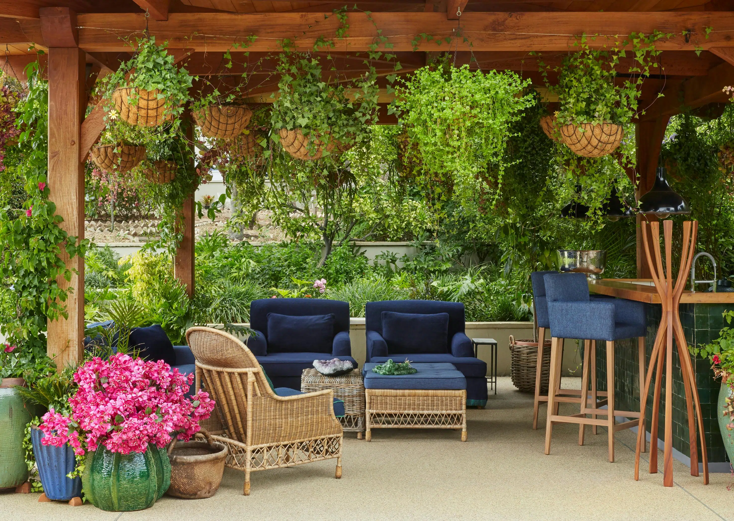 An outdoor entertaining area featuring rattan furniture, surrounded by greenery, with a bar area to the left.