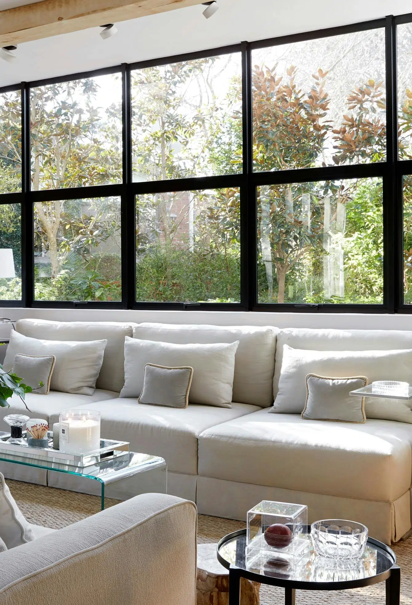 A modern living room with a glass coffee table, a white sofa, and large windows with a view of the greenery outside.