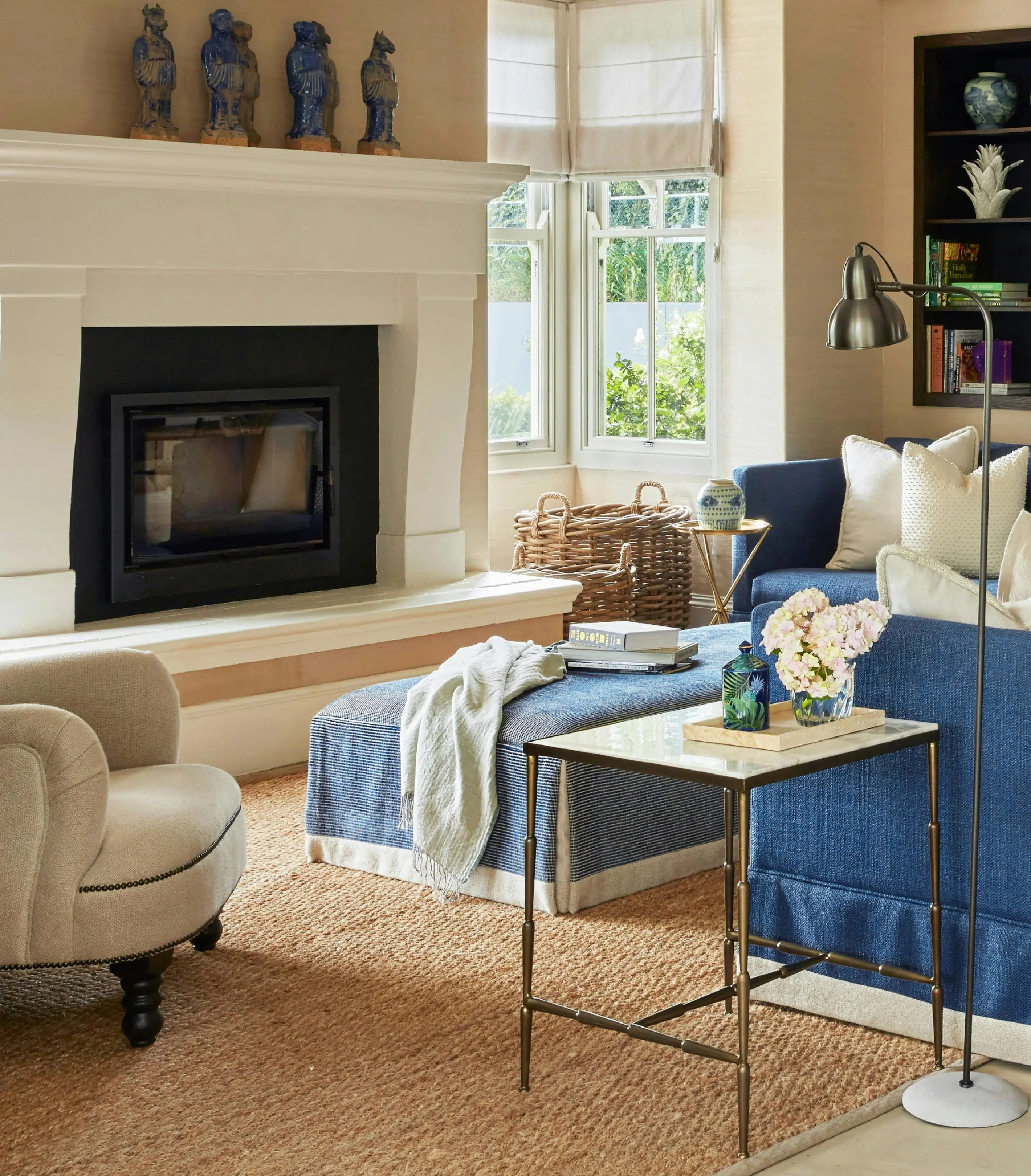 A living room is furnished in blues, creams and white, with chairs and an ottoman visible in front of a large fireplace with a mantlepiece holding blue ceramic figures.