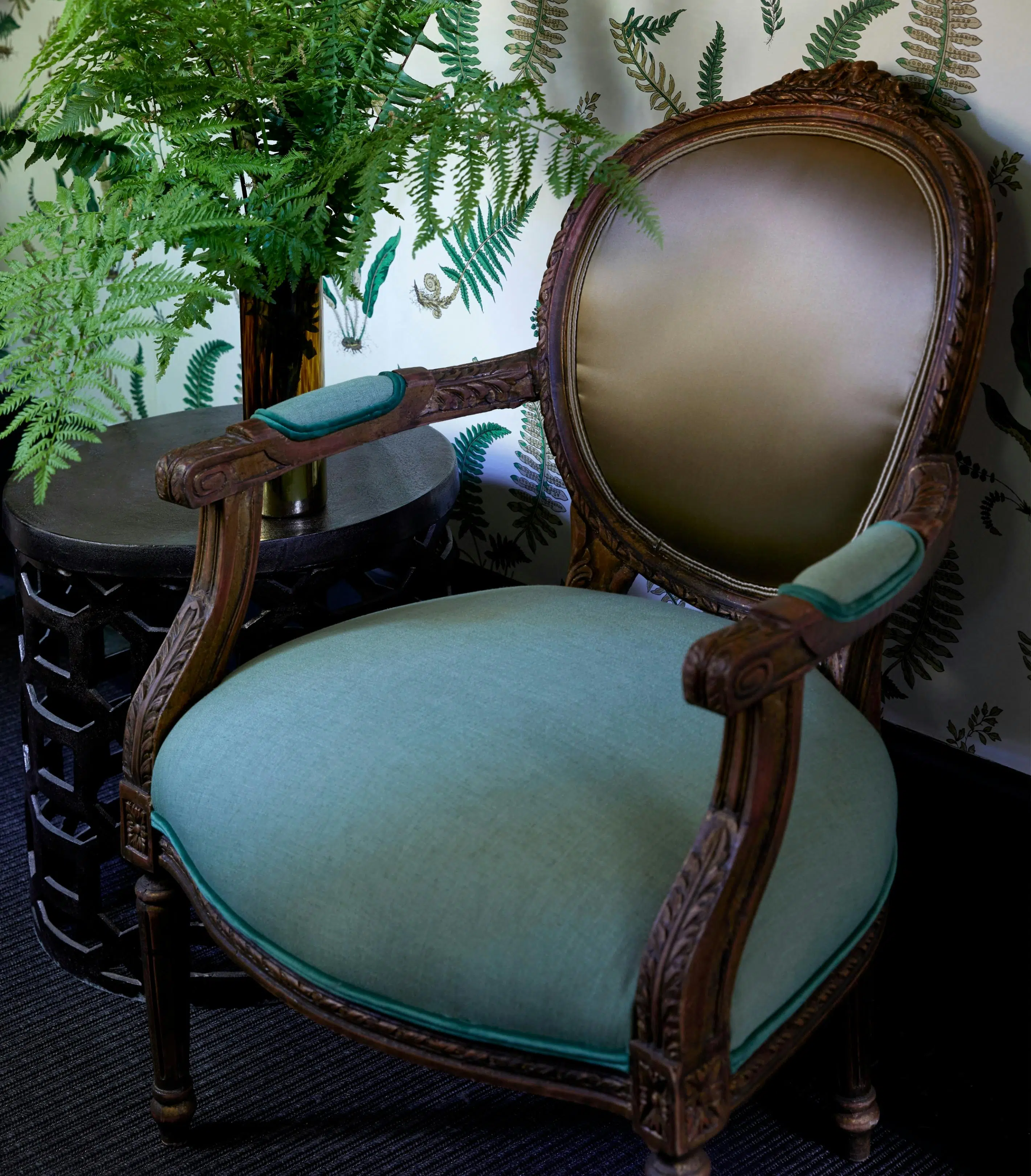 A Louis XV-style chair with a dark wooden frame has its back upholstered in a glossy, brown fabric while its seat and arm rests are upholstered in a colour close to duck egg blue. Wallpaper behind it is illustrated with ferns, and a vase of ferns sits beside it on a side table