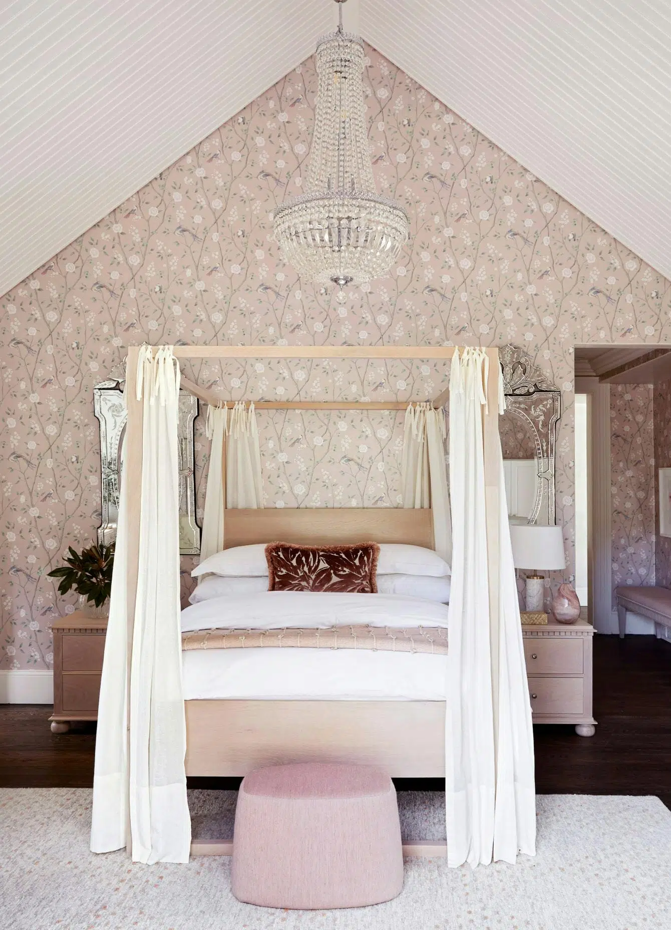 A sophisticated bedroom with a 4-poster bed in the centre, patterned wallpaper on the back wall and a pouffe just in front of the bed on the rug.