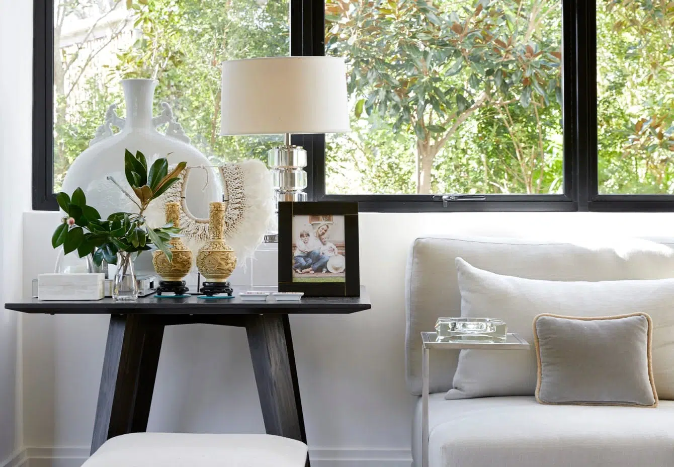 Close-up of a living room with two side tables with decorative accents on top, a window with a view of a tree outside, and the corner of a white sofa.