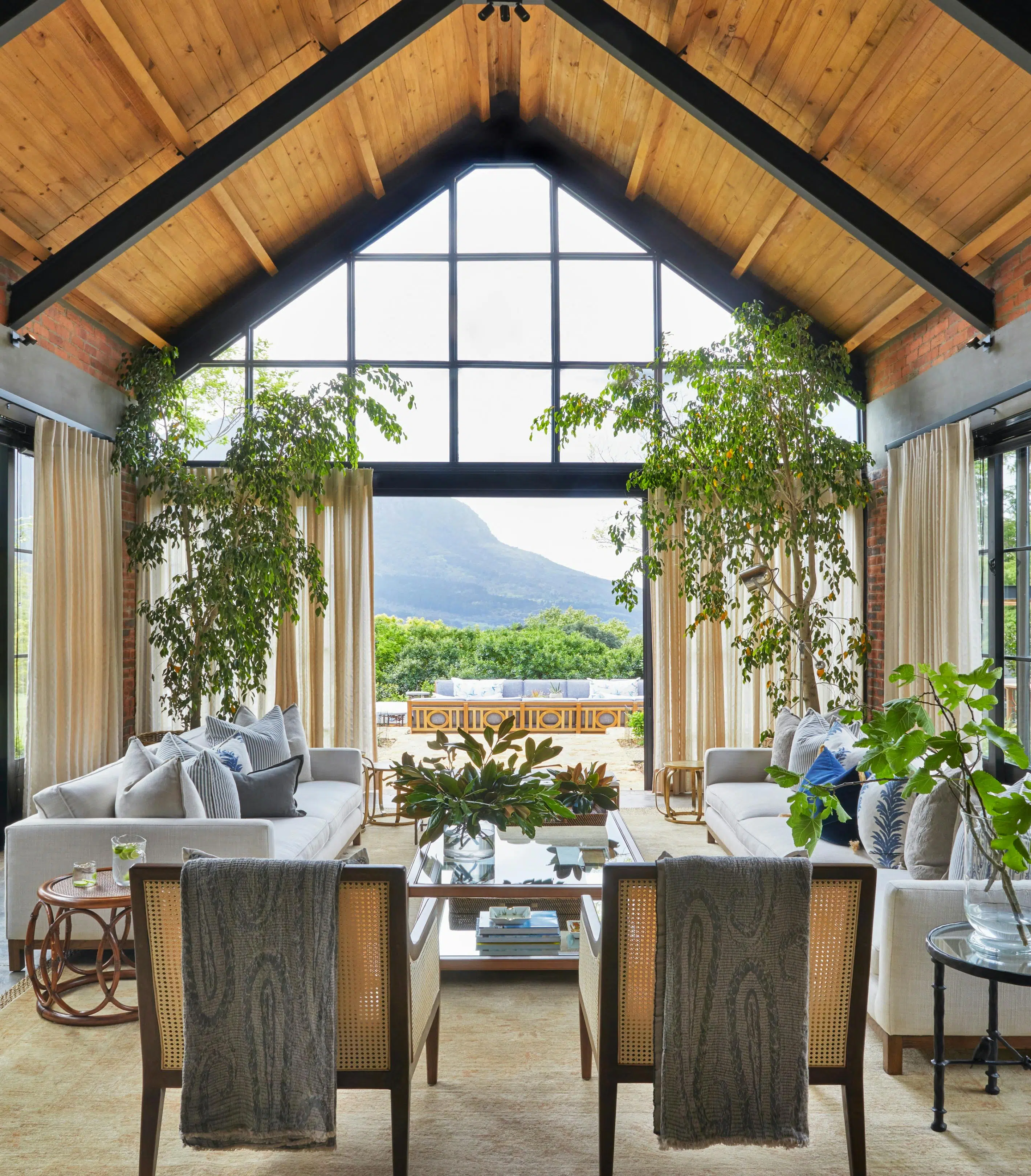 A living room with an a-frame roof, two occasional chairs and two sofas in the foreground with large plants that frame the door to the outside area in the background