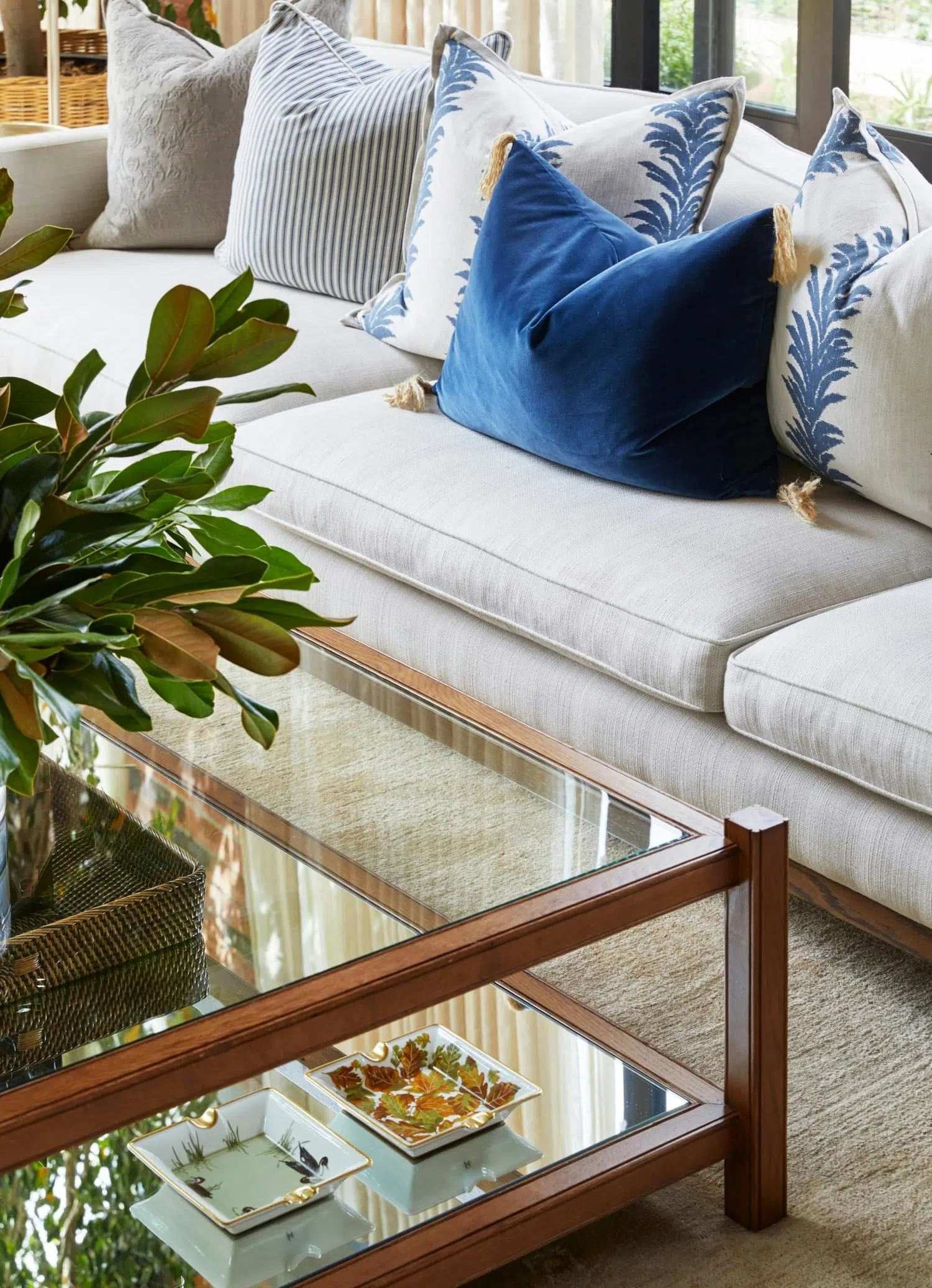 A wooden-framed coffee table with a mirrored base and clear glass table top holds ornate ashtrays on its lower levels and what appears to be a vase of greenery on its top. A neutral coloured couch is visible, holding blue and blue-patterned cushions.