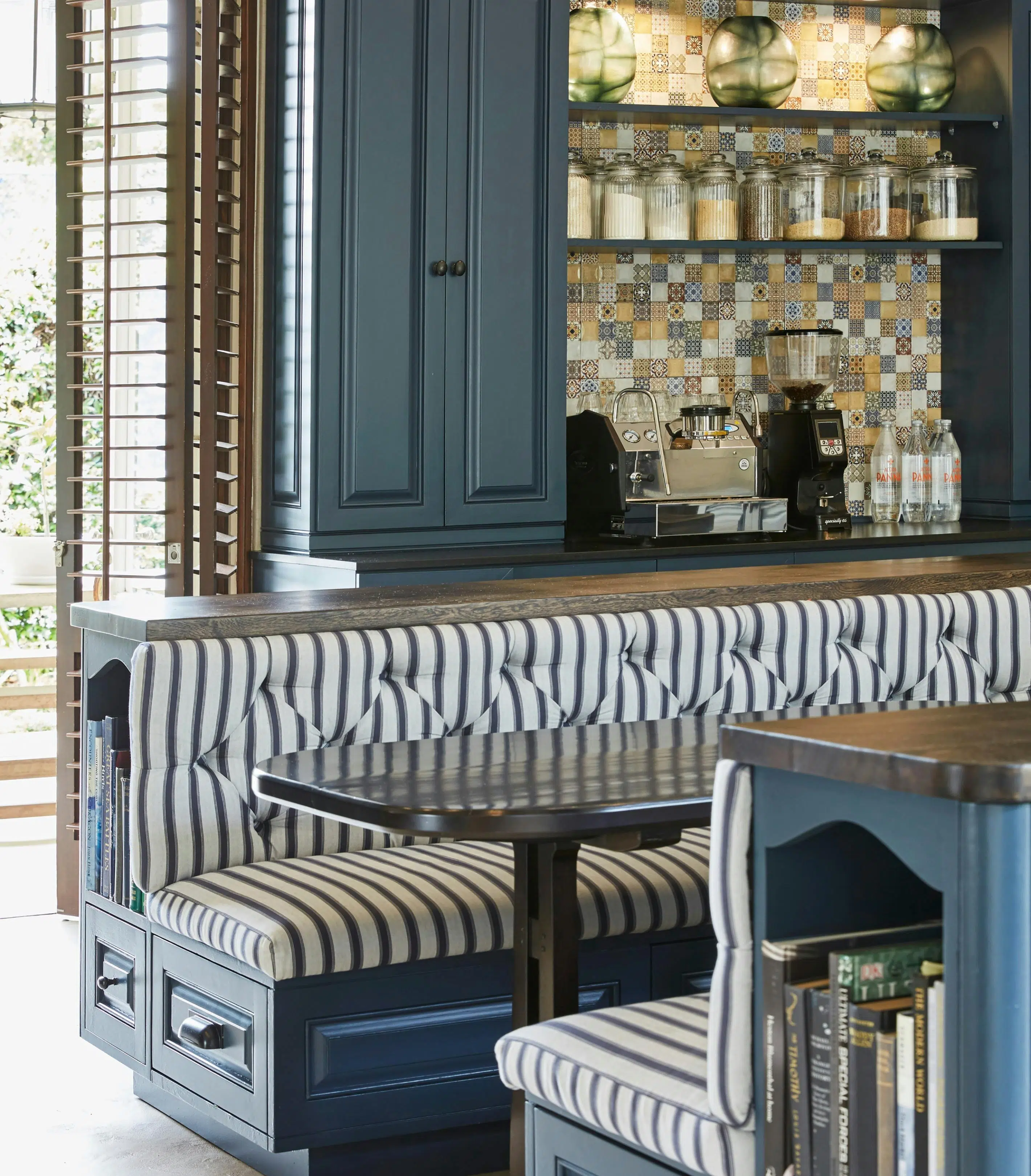 A breakfast nook, in dark blue-painted wood with built-in bookshelves and drawers, can be seen in front of a tall kitchen cupboard in the same treatment. A tiled section of the wall separates an area for a coffee machine, bean grinder, and shelves holding dry goods.