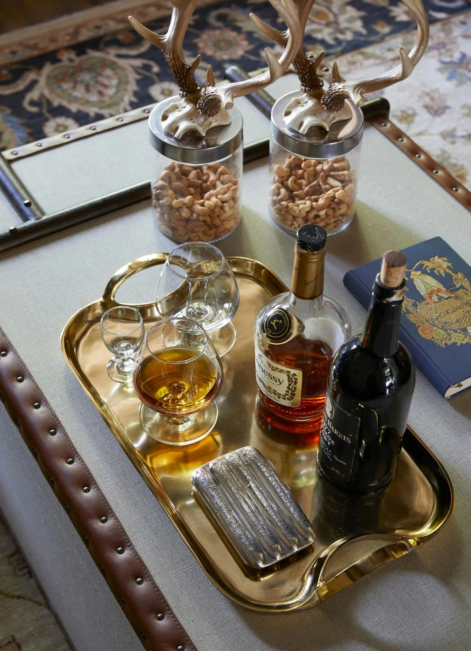 A bottle of Hennessy and a bottle of port sit on a gold tray with some glasses and what appears to be a cigarette box. Two jars contain cashew nuts, and in the background a patterned carpet is visible.