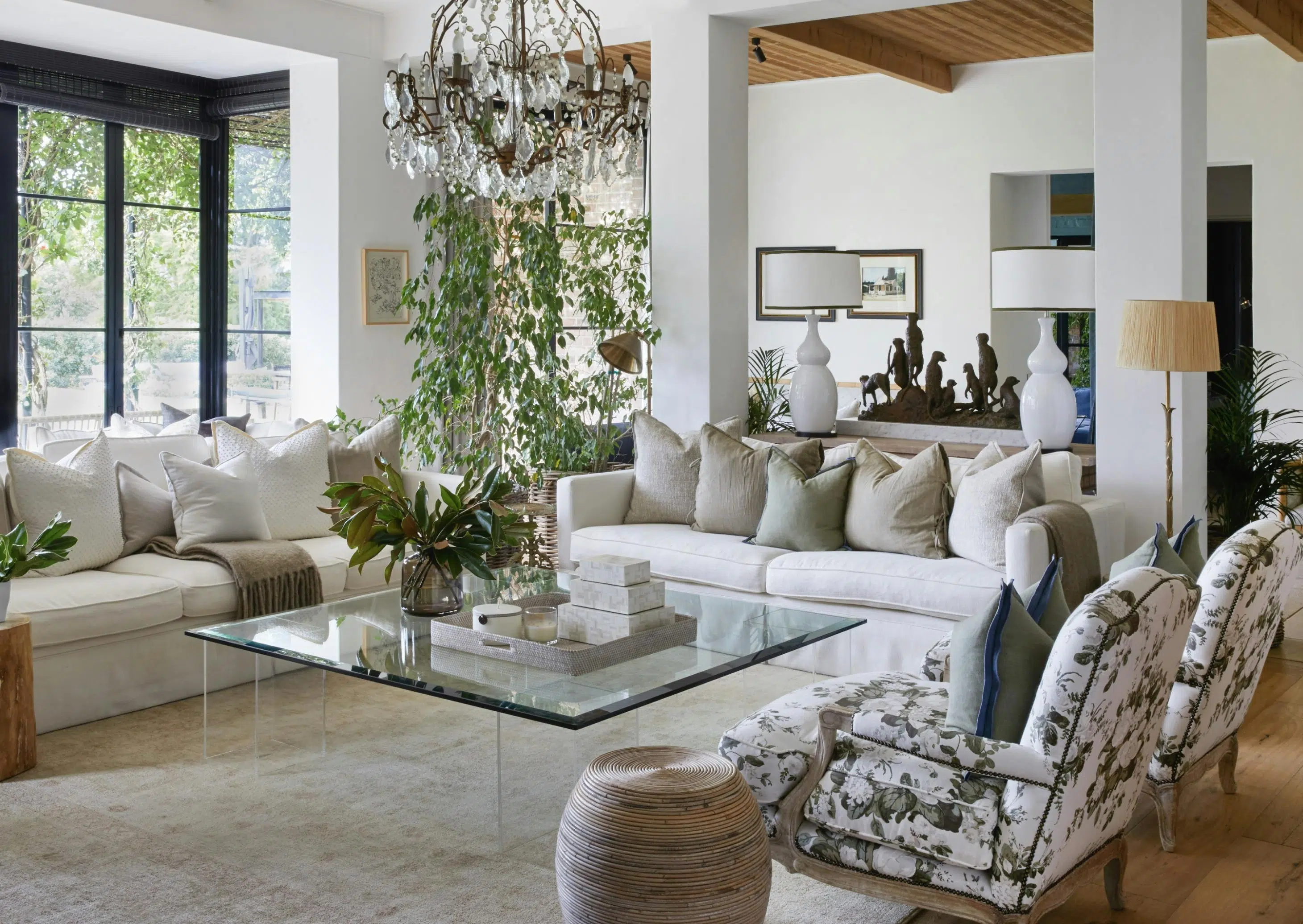 A sitting area features two couches, upholstered chairs, and a glass coffee table holding a tray of square objects and a glass vase of greenery. A leafy plant, a pair of white lamps, and a sculpture of a group of meerkats are visible in the background. A chandelier hangs above.