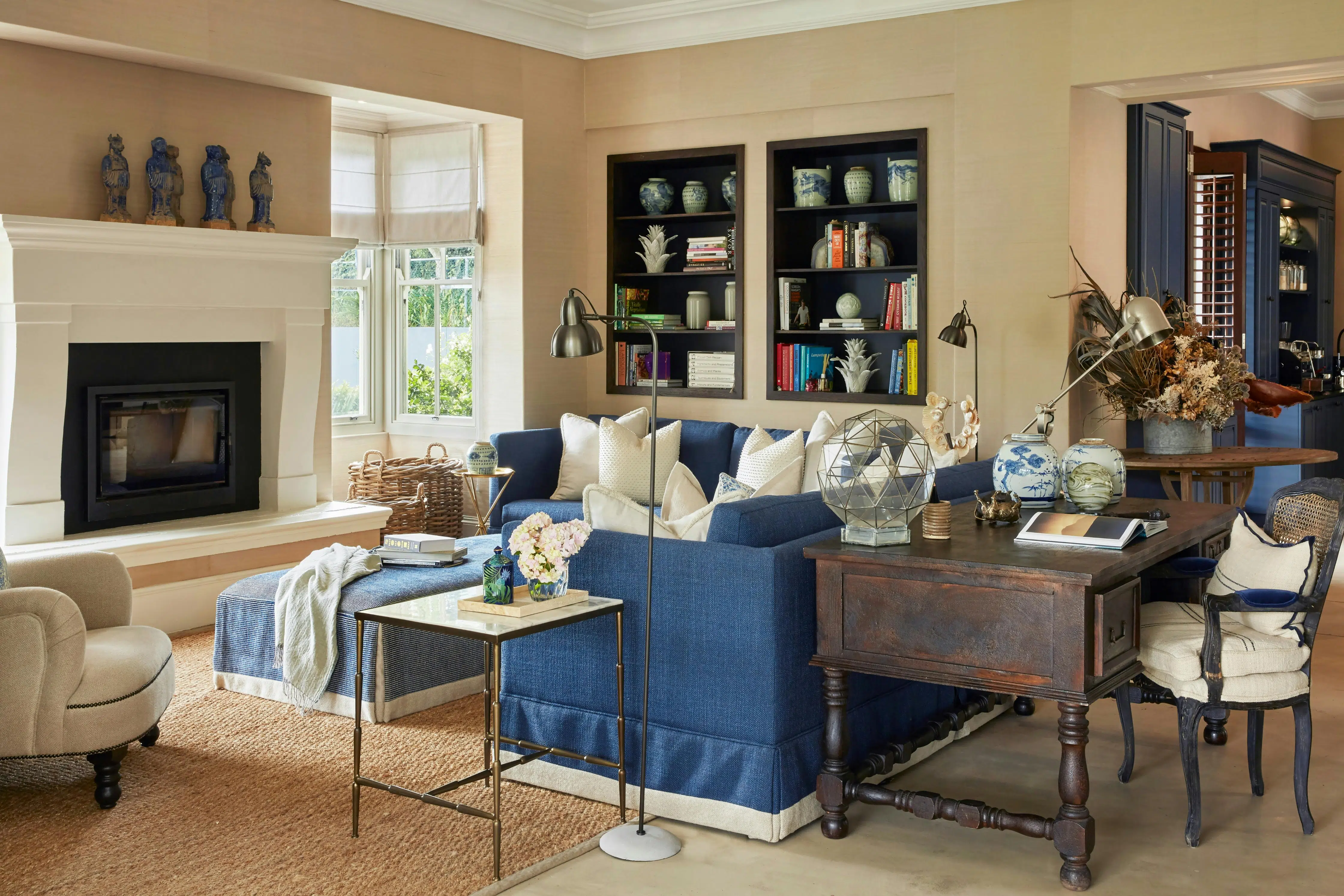 A comfortably furnished living room features couches upholstered in blue, a heavy wooden writing desk, bookshelves and a large, simple fireplace.