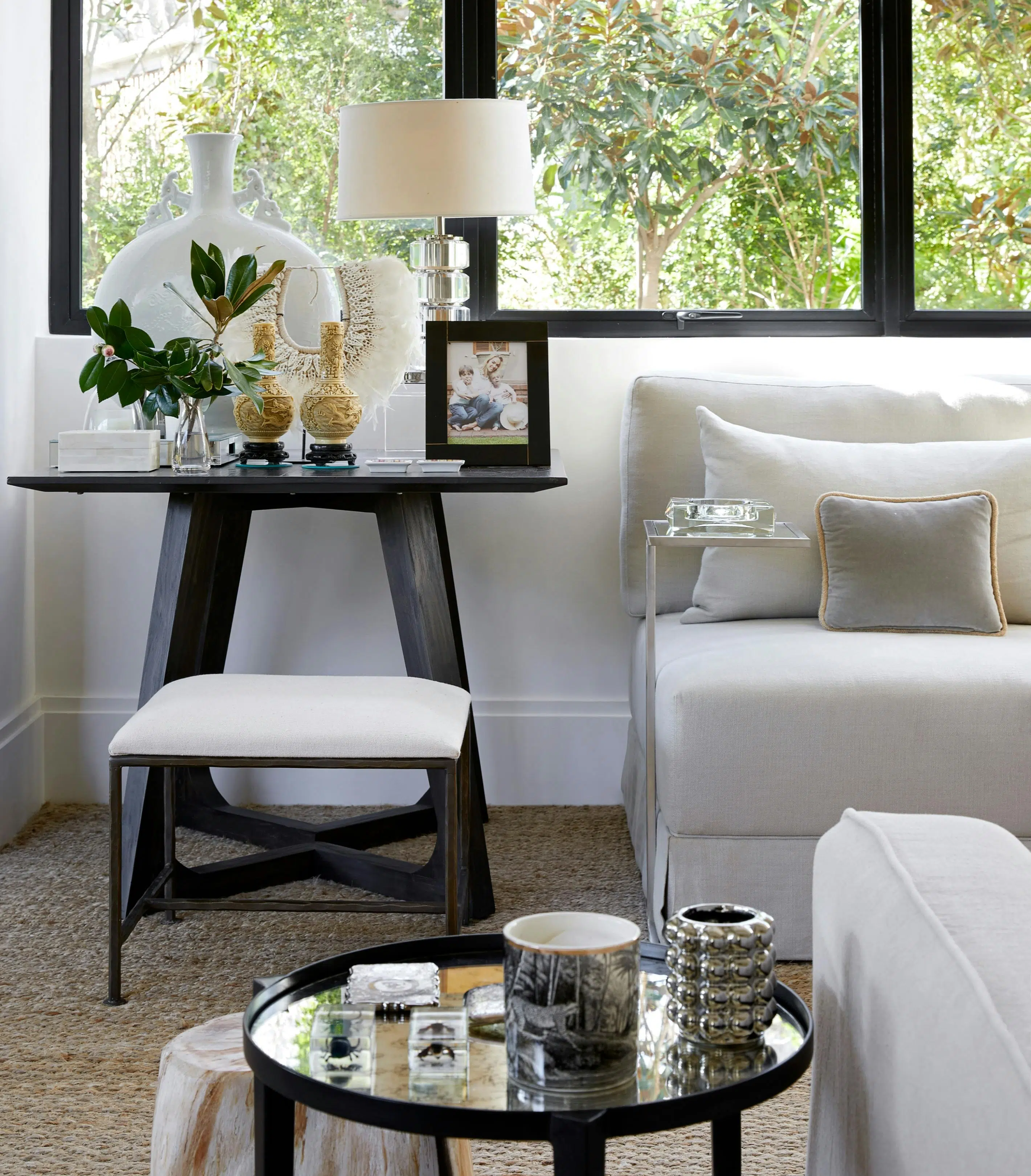 Close-up of a living room with two side tables with decorative accents on top, a window with a view of a tree outside, and the corner of a white sofa.