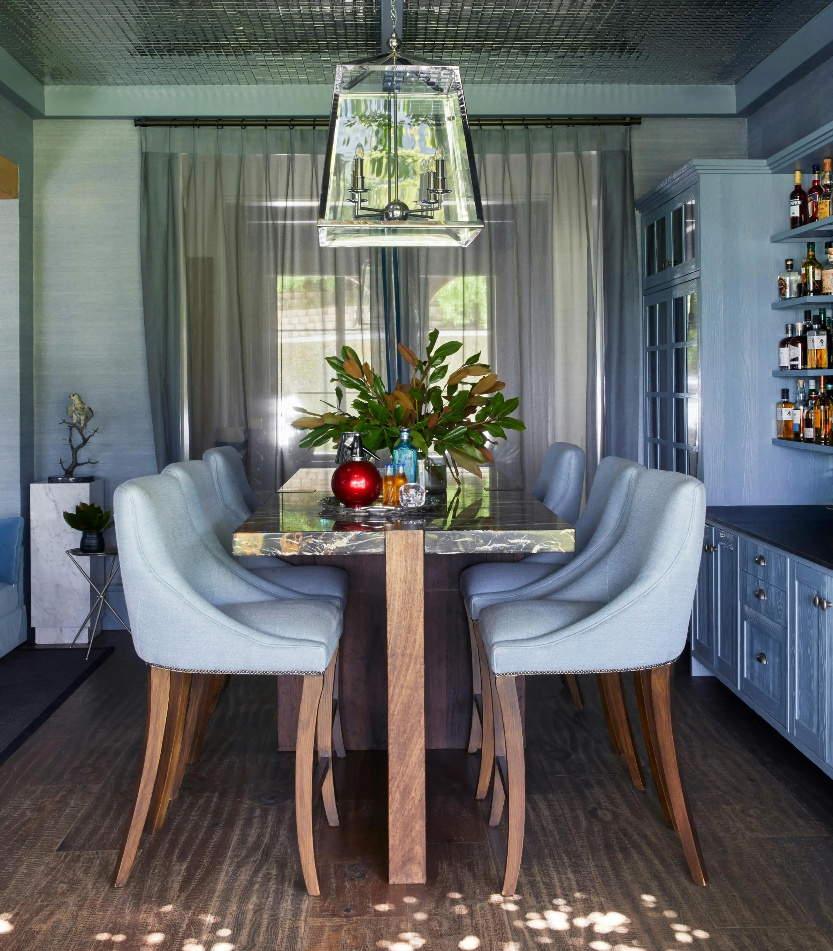 A bar counter has blue-upholstered chairs arranged in front of and behind it. Shelves holding the bar's bottles of liquor can be seen to one side, and a bold, glass pendant light hangs above the countertop