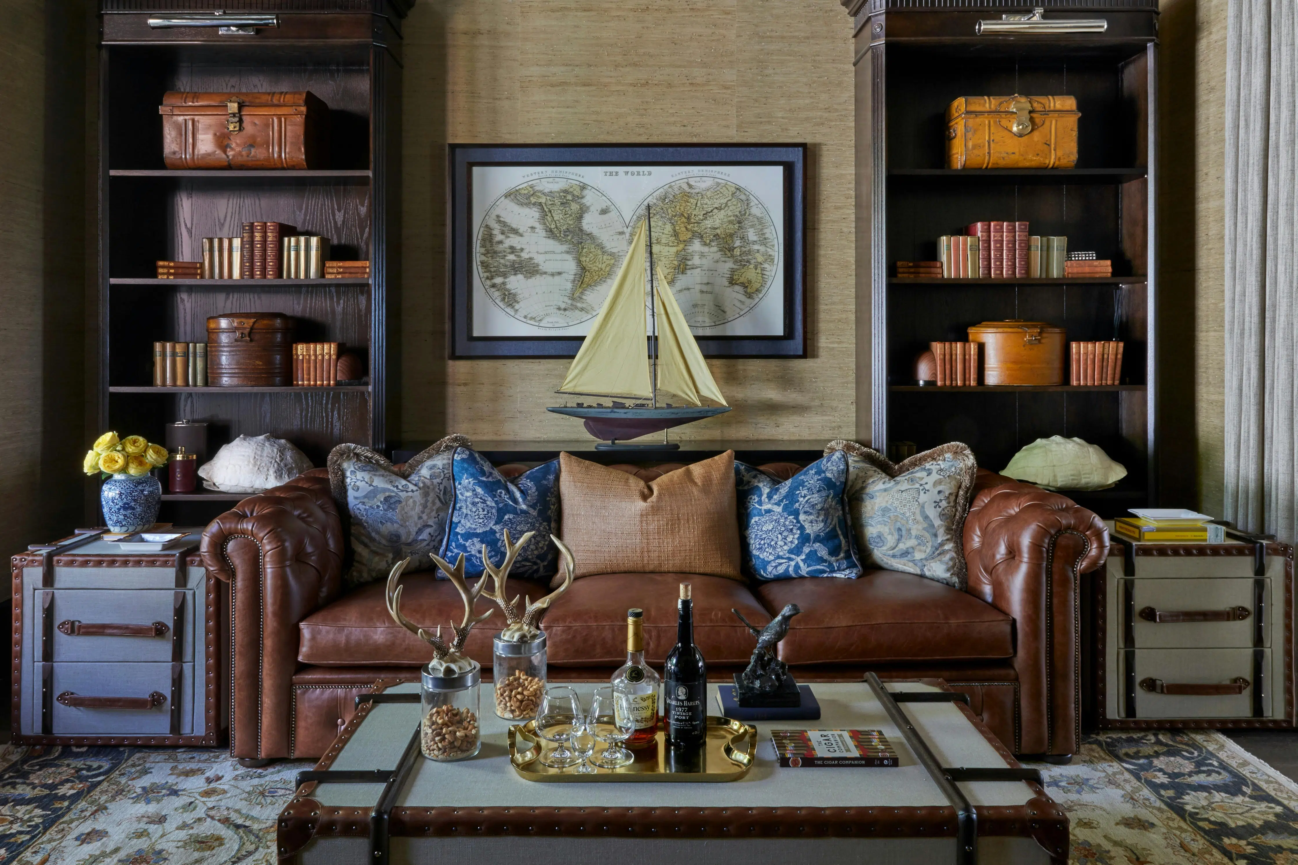 A brown leather sofa sits in the centre of a room, flanked by side tables bound in leather and fabric, much like luggage. Behind the couch are bookshelves and a framed, vintage map. In front of it is a coffee table in a similar style to the side tables, holding a drinks tray and two jars adorned with imitation antlers and holding cashew nuts