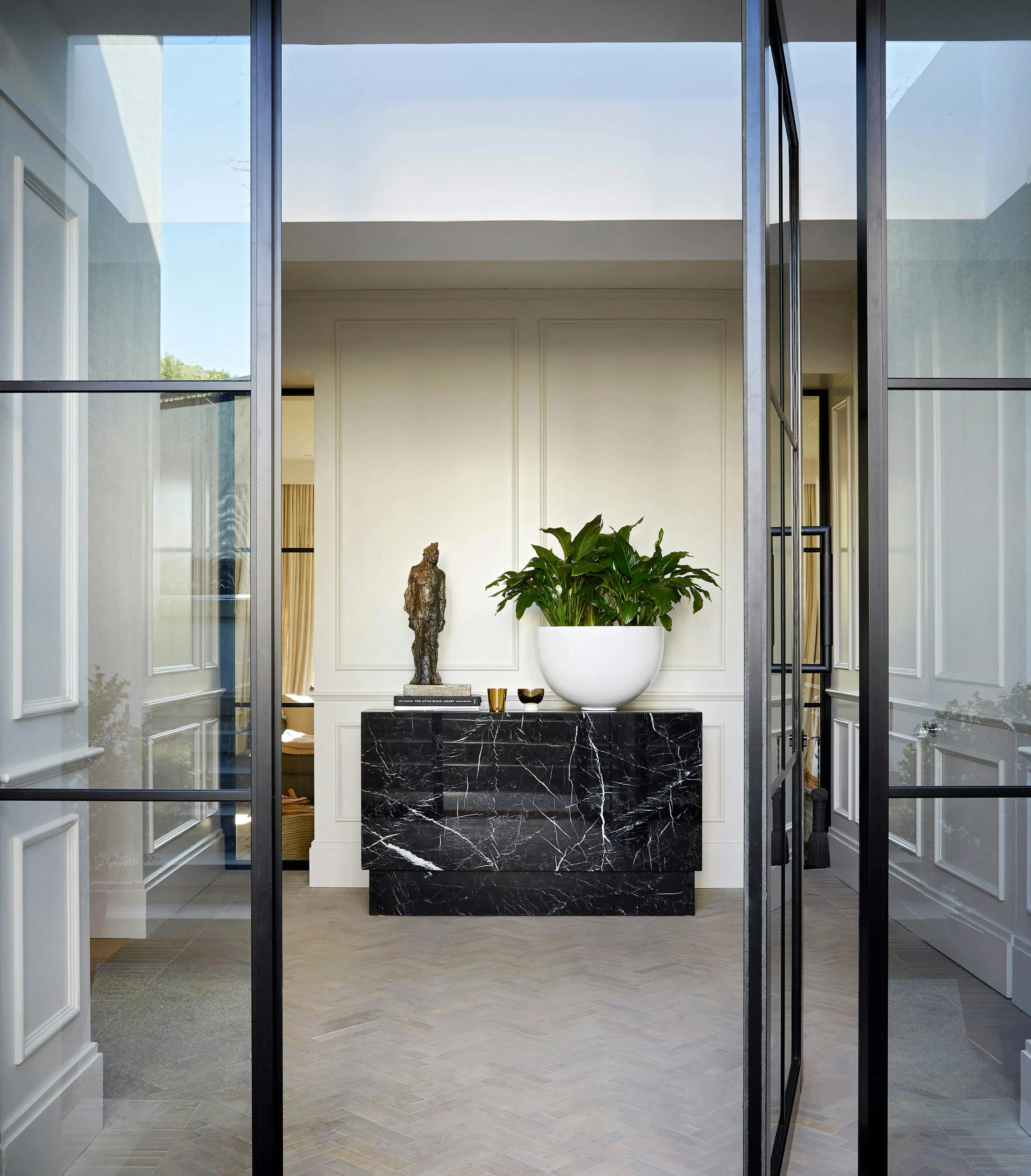 View of a front entrance to a home with with a large potted plant and a sculpture of a man on top of a marble console table in the centre.